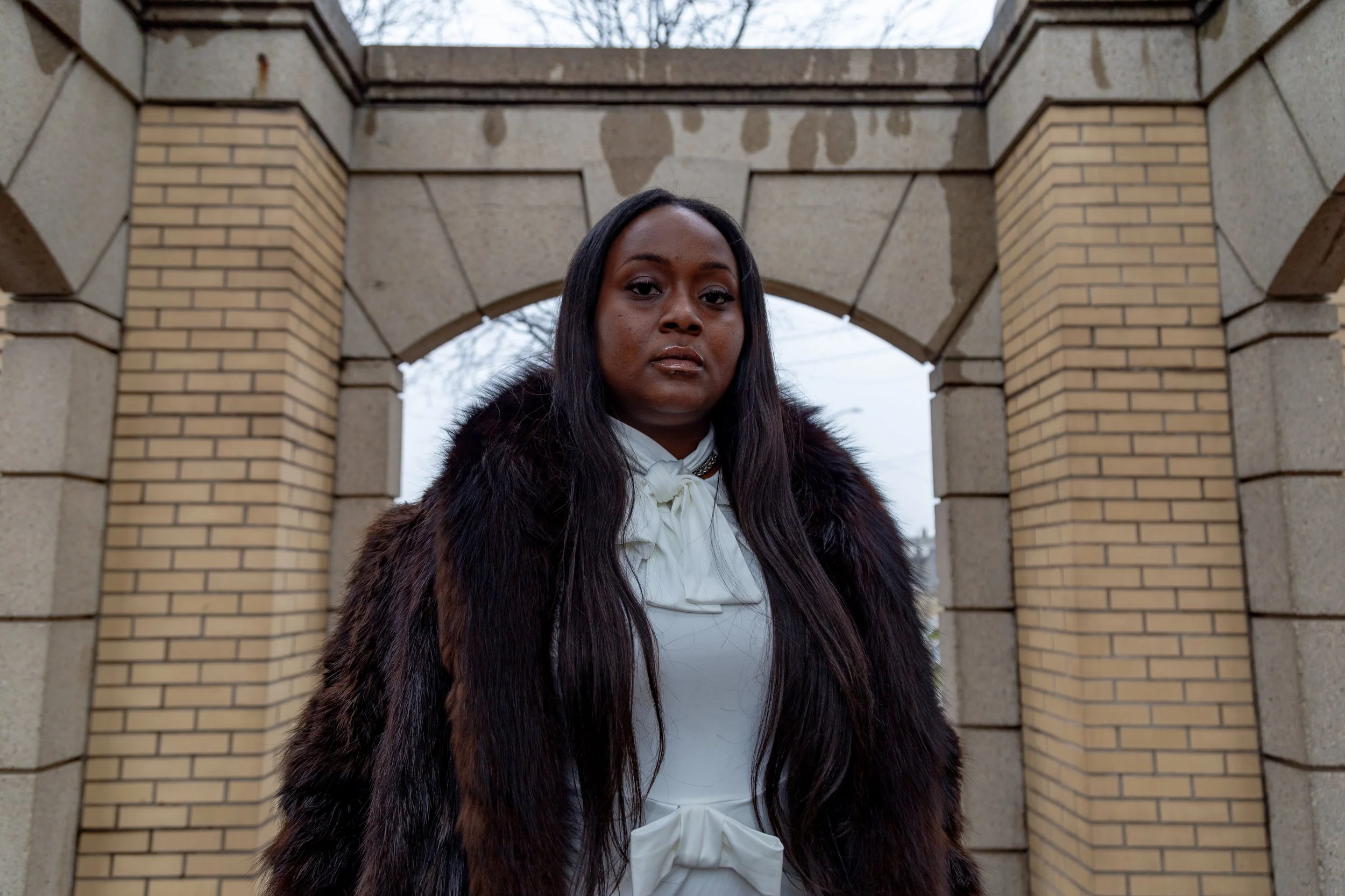 A woman with long dark hair wearing a dark fur coat and white blouse stands outdoors in front of a stone archway, with an overcast sky in the background.