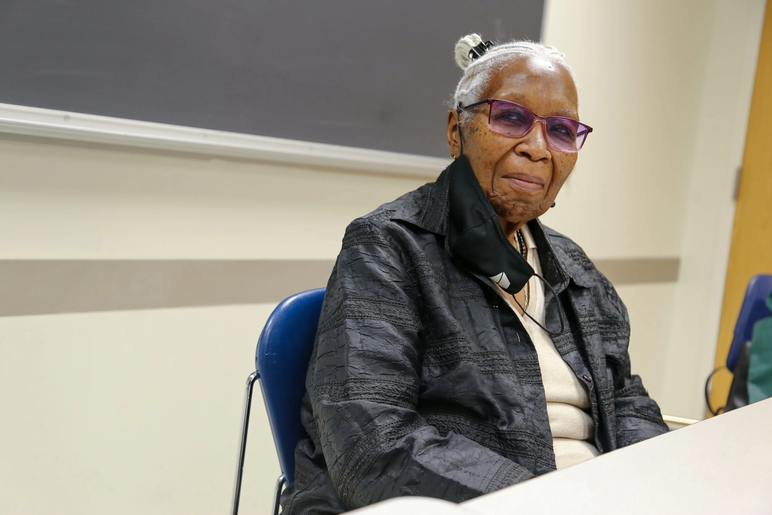 An elderly woman with gray hair tied back, wearing glasses, a black jacket, light-colored shirt, and a face mask pulled down to her chin, sitting in a classroom or meeting room with a blackboard behind her.