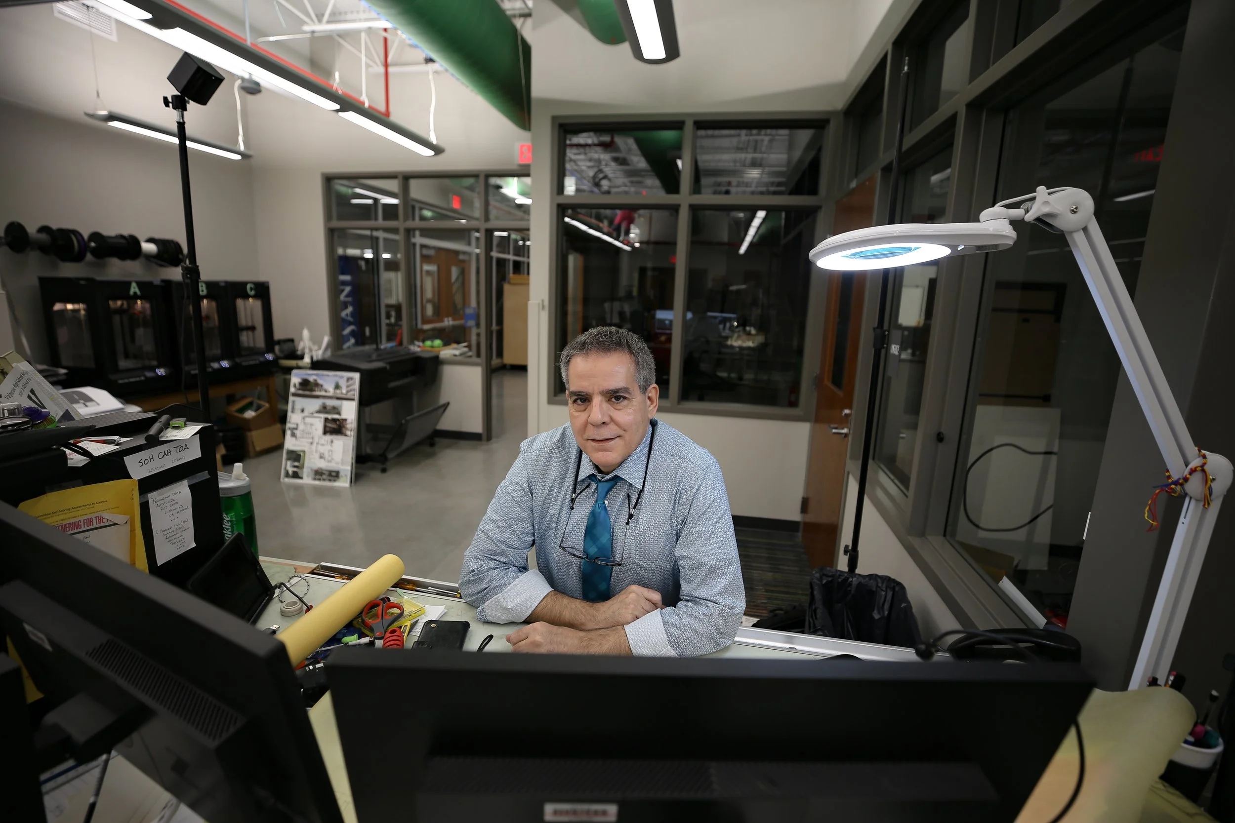 Man in a lab coat and blue tie sitting at a desk in an office with computers, equipment, and large windows.
