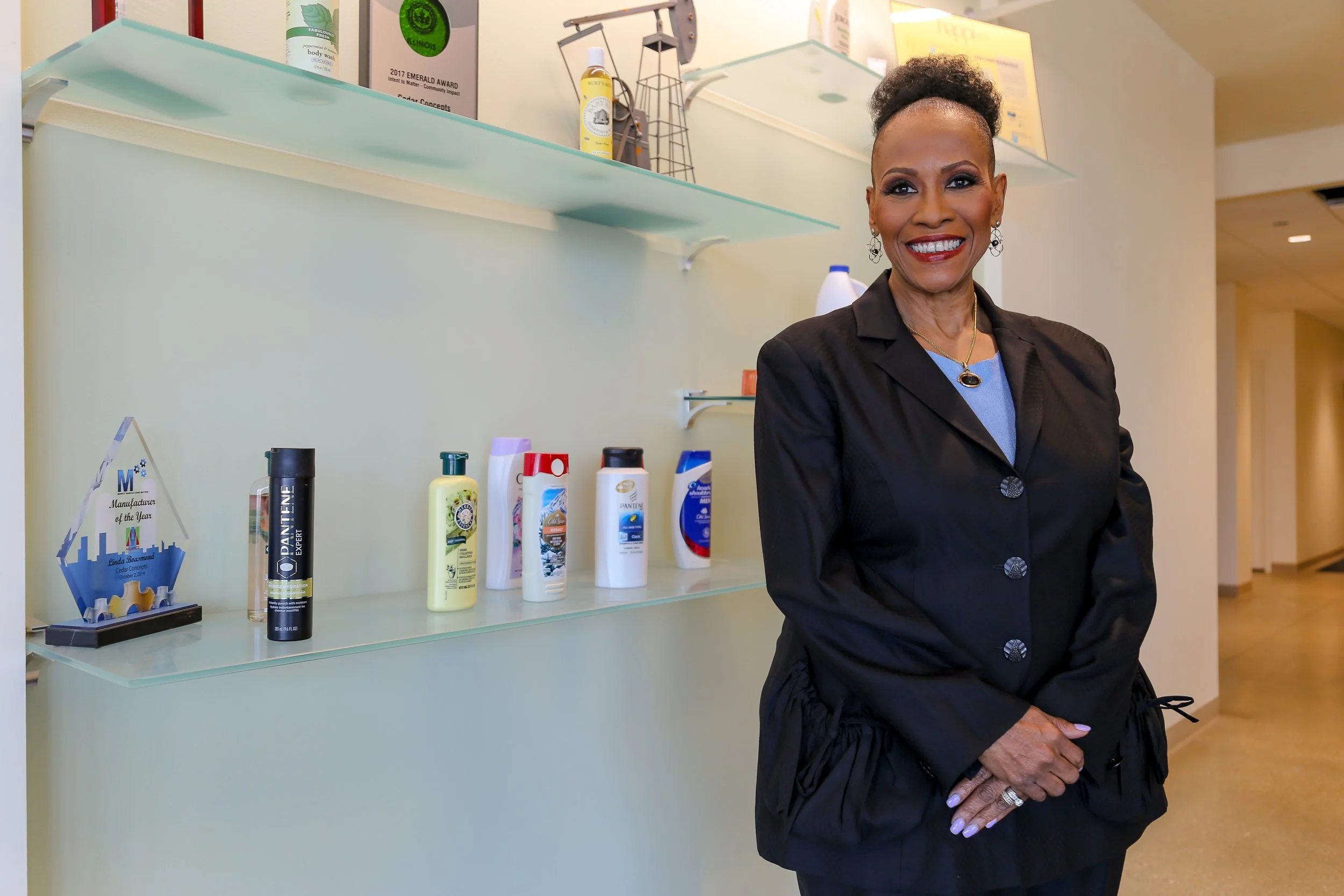 A smiling woman in a black blazer standing in front of a shelf with various personal care products in an indoor setting.