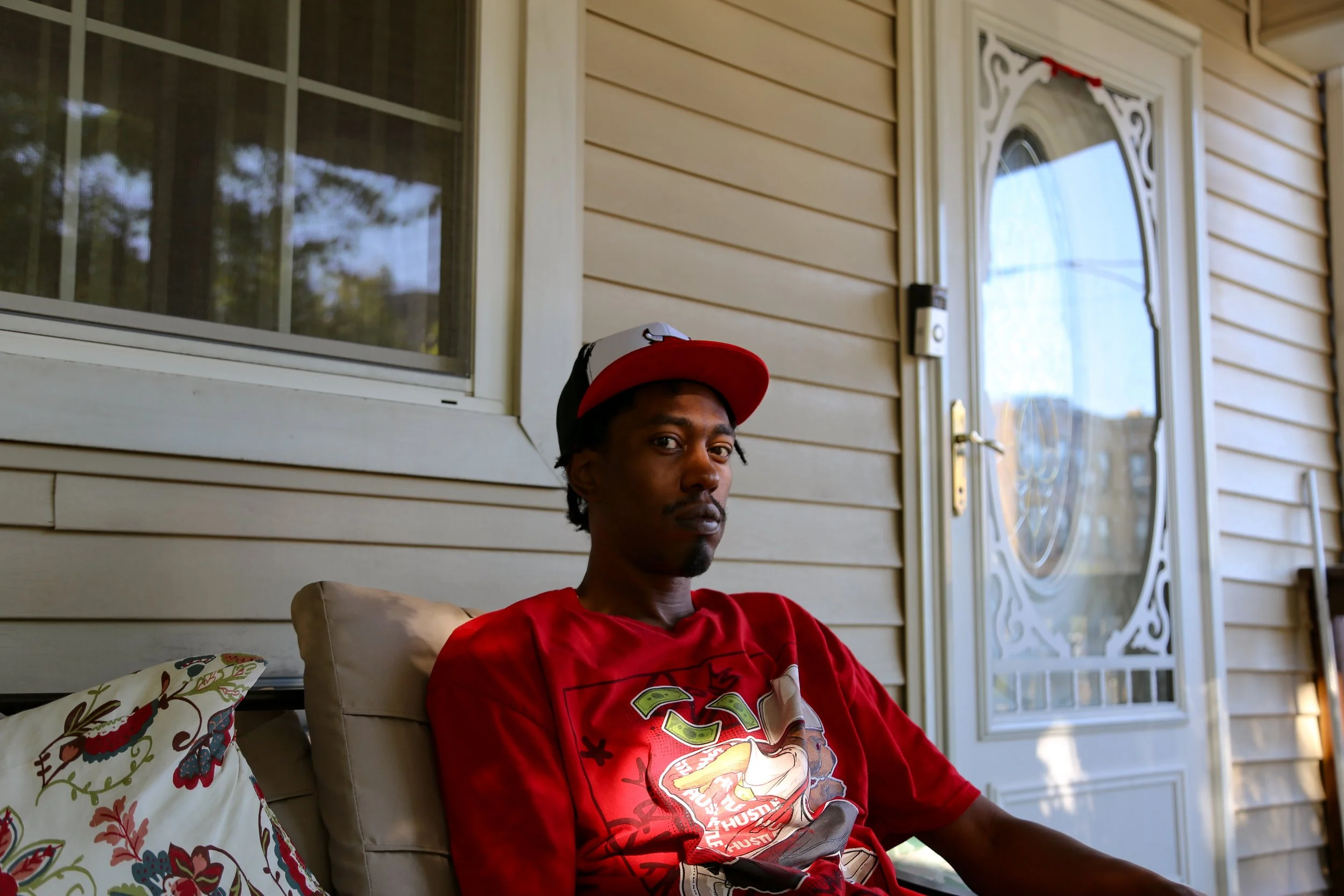 A young man wearing a red T-shirt and a black, white, and red baseball cap sitting on a porch with beige siding, a decorative glass door, and an outdoor cushion with a floral pattern.