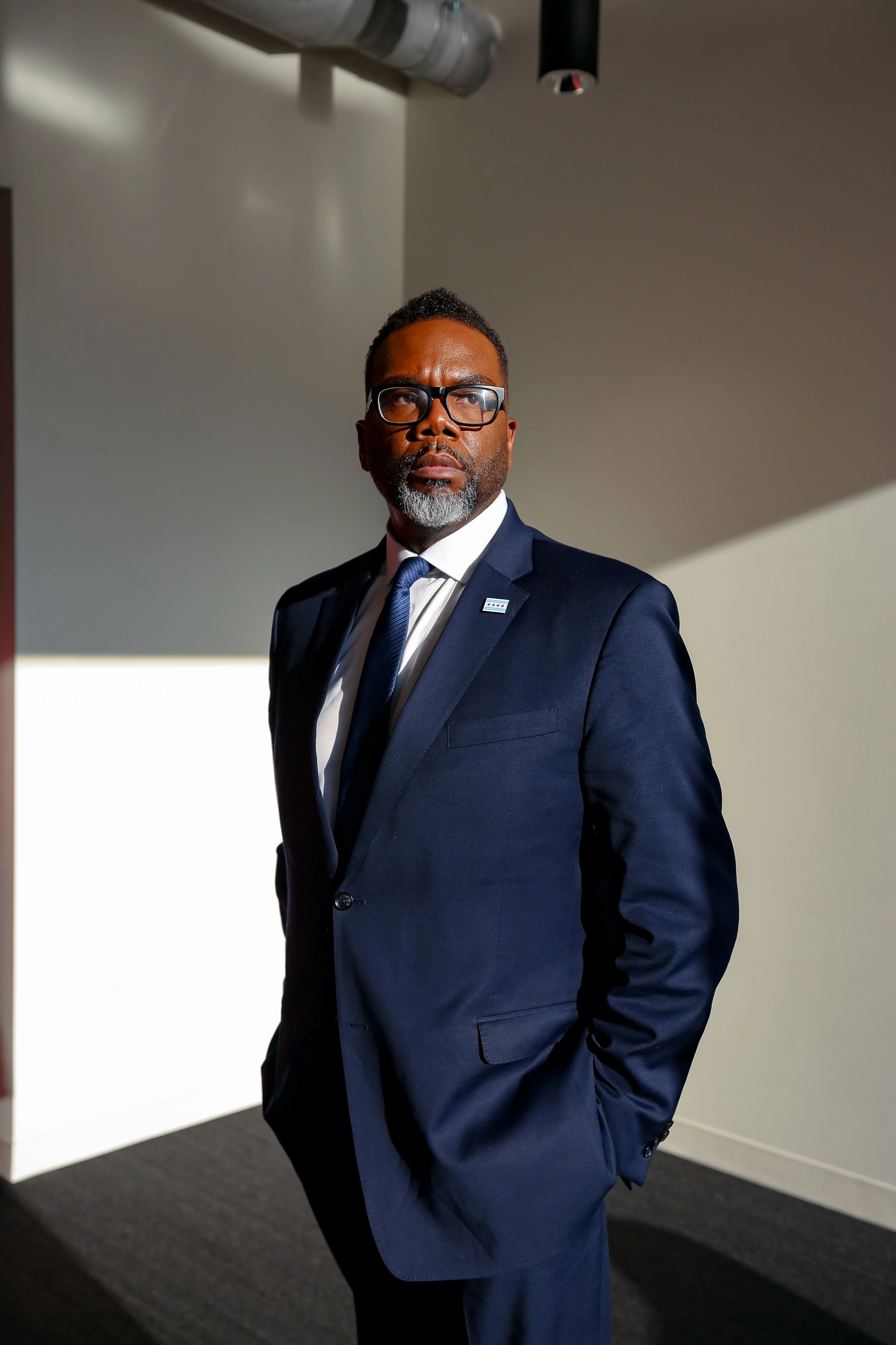 A man in a navy blue suit and tie standing indoors with one hand in his pocket, wearing glasses, with a serious expression, under a ceiling light and a silver ventilation pipe.
