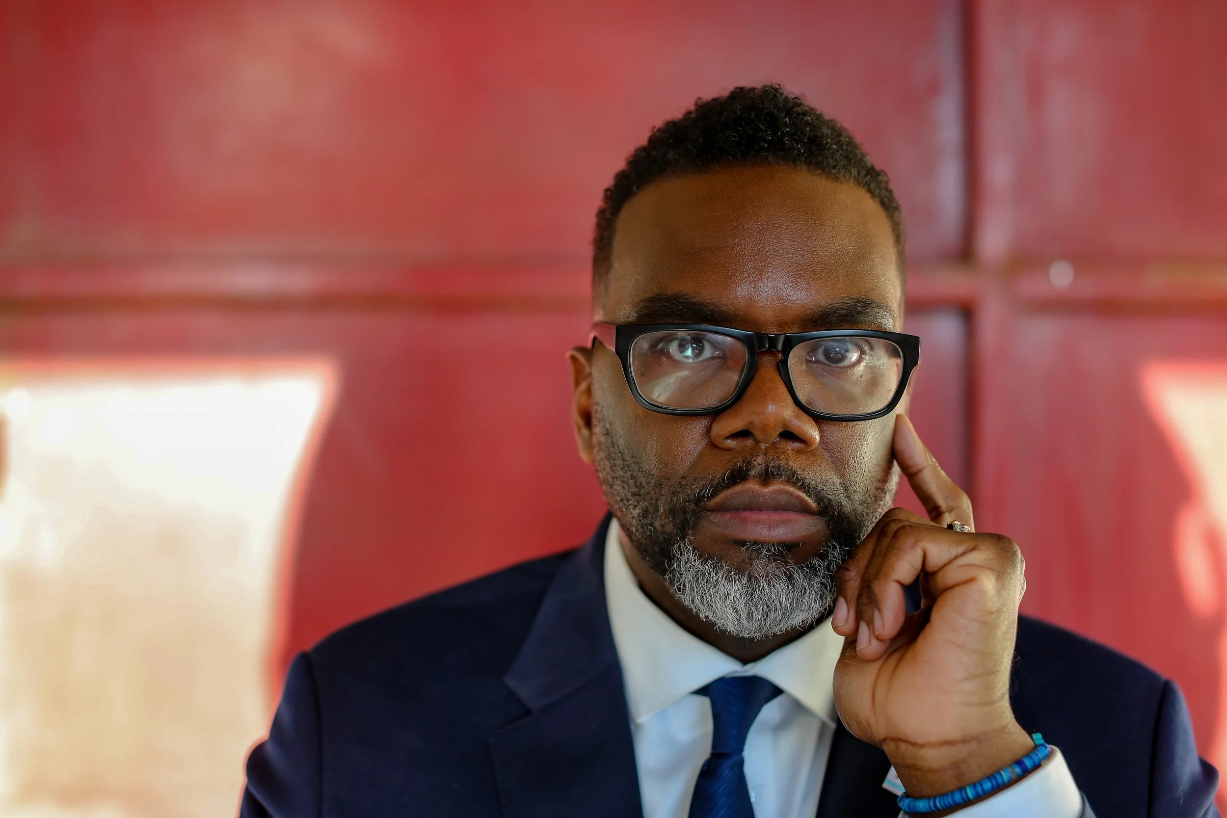 A middle-aged man with black hair and a beard, wearing glasses, a dark blue suit, white shirt, and blue tie, resting his chin on his hand in front of a red background.