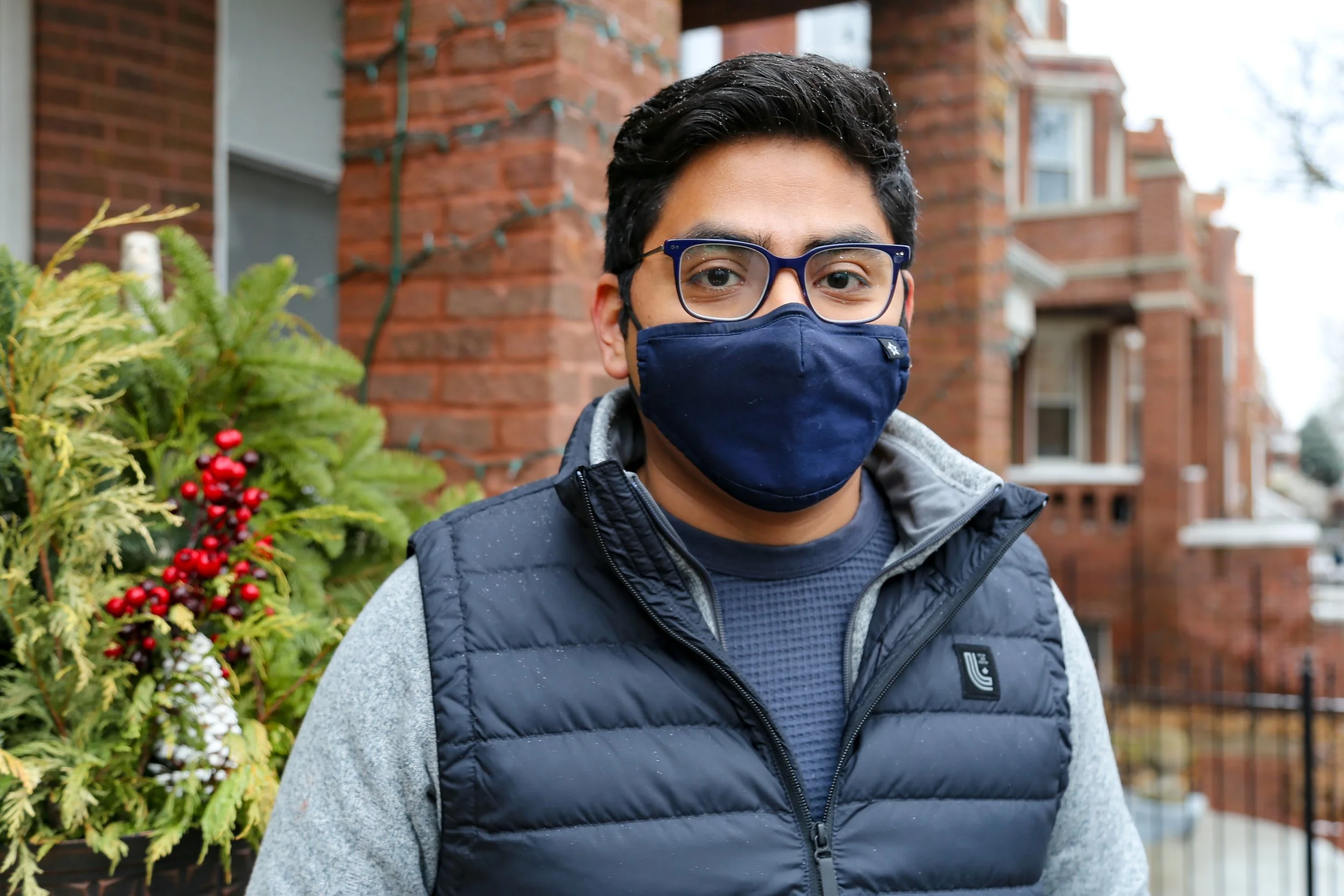 A man wearing glasses and a dark blue face mask standing outdoors in front of a brick building and greenery with red berries.