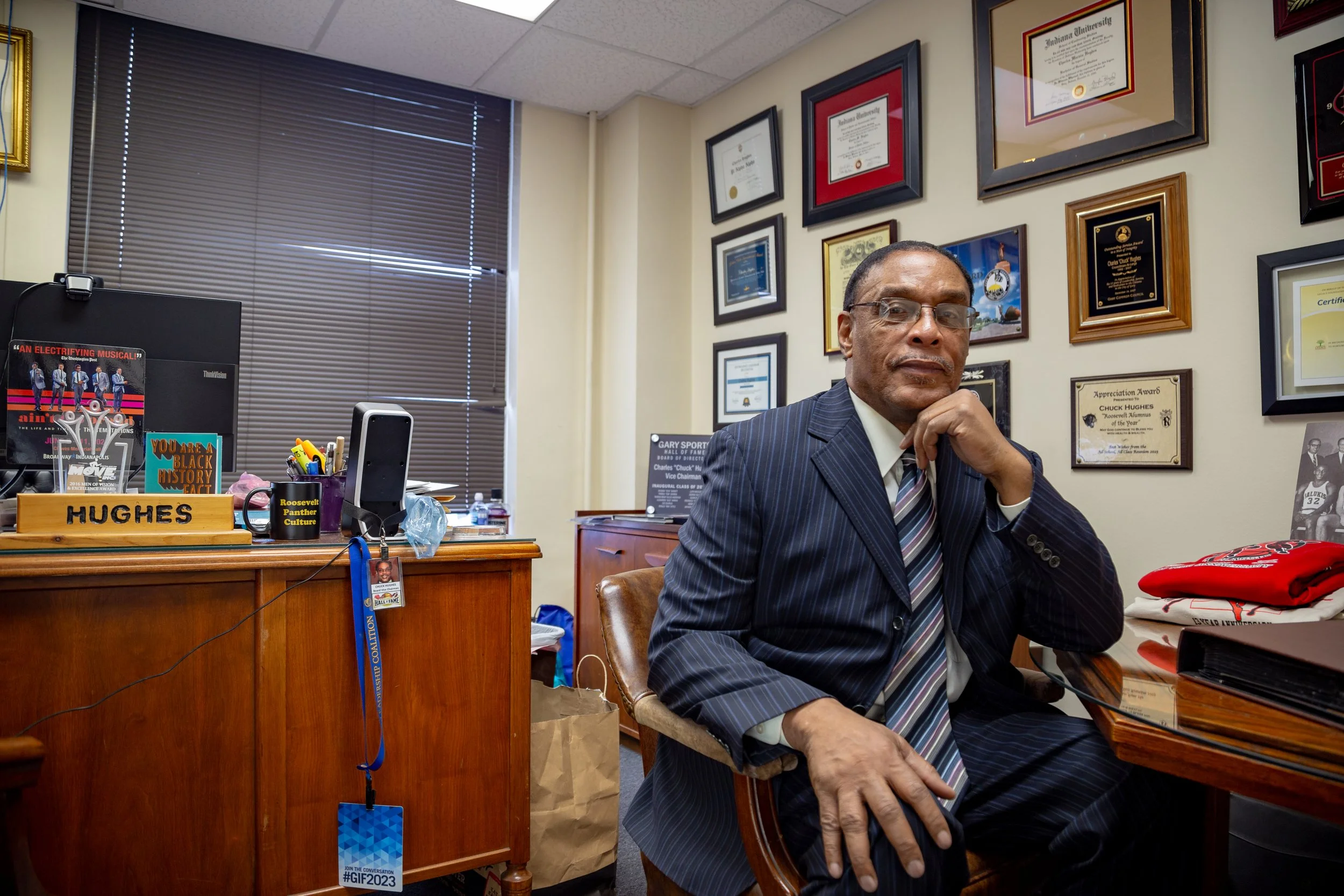 A man in a dark pinstripe suit sitting at a desk in an office decorated with plaques and certificates on the wall.