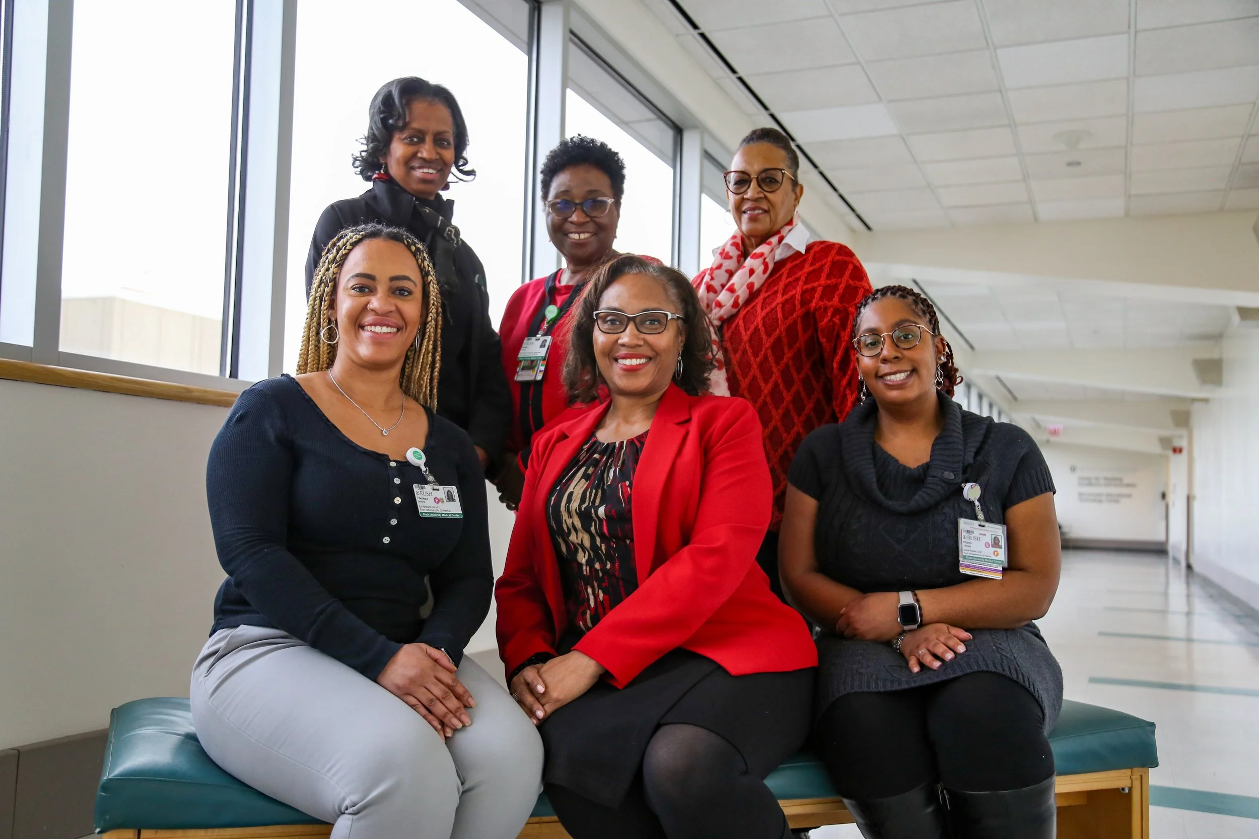 Group of seven women sitting and standing in a hospital corridor with large windows, wearing professional attire and identification badges, smiling at the camera.