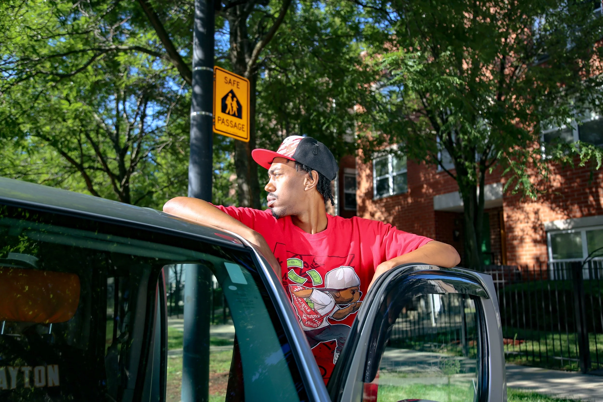 Young man wearing a red shirt and baseball cap leaning on a car window, standing outside in a green area with trees and a brick building in the background.