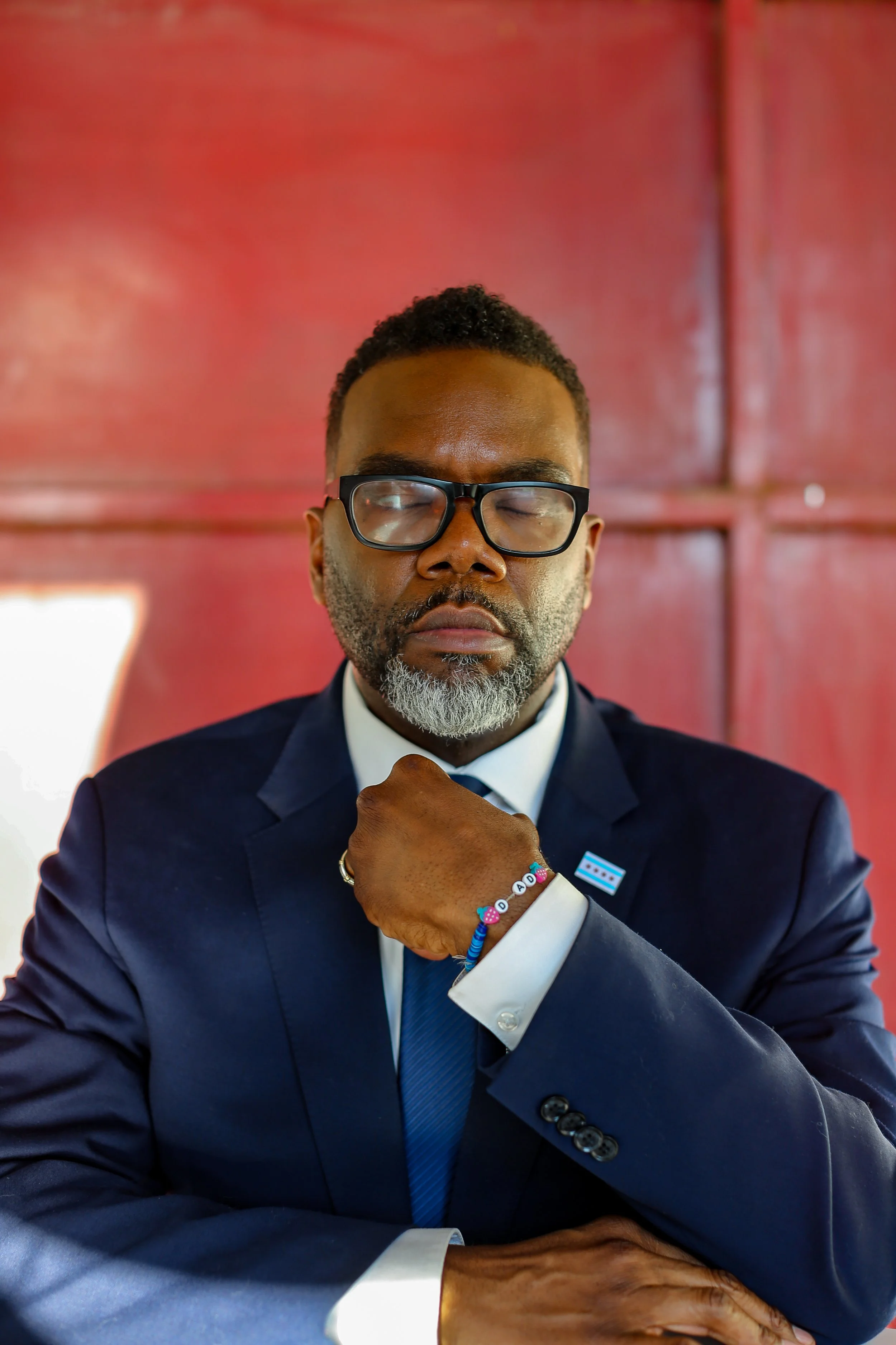 A man wearing a navy blue suit, white shirt, and blue tie is sitting with his eyes closed, resting his chin on his fist, showing a bracelet on his wrist and a pin on his lapel. The man has black glasses, short black hair, and gray beard, and is in fr