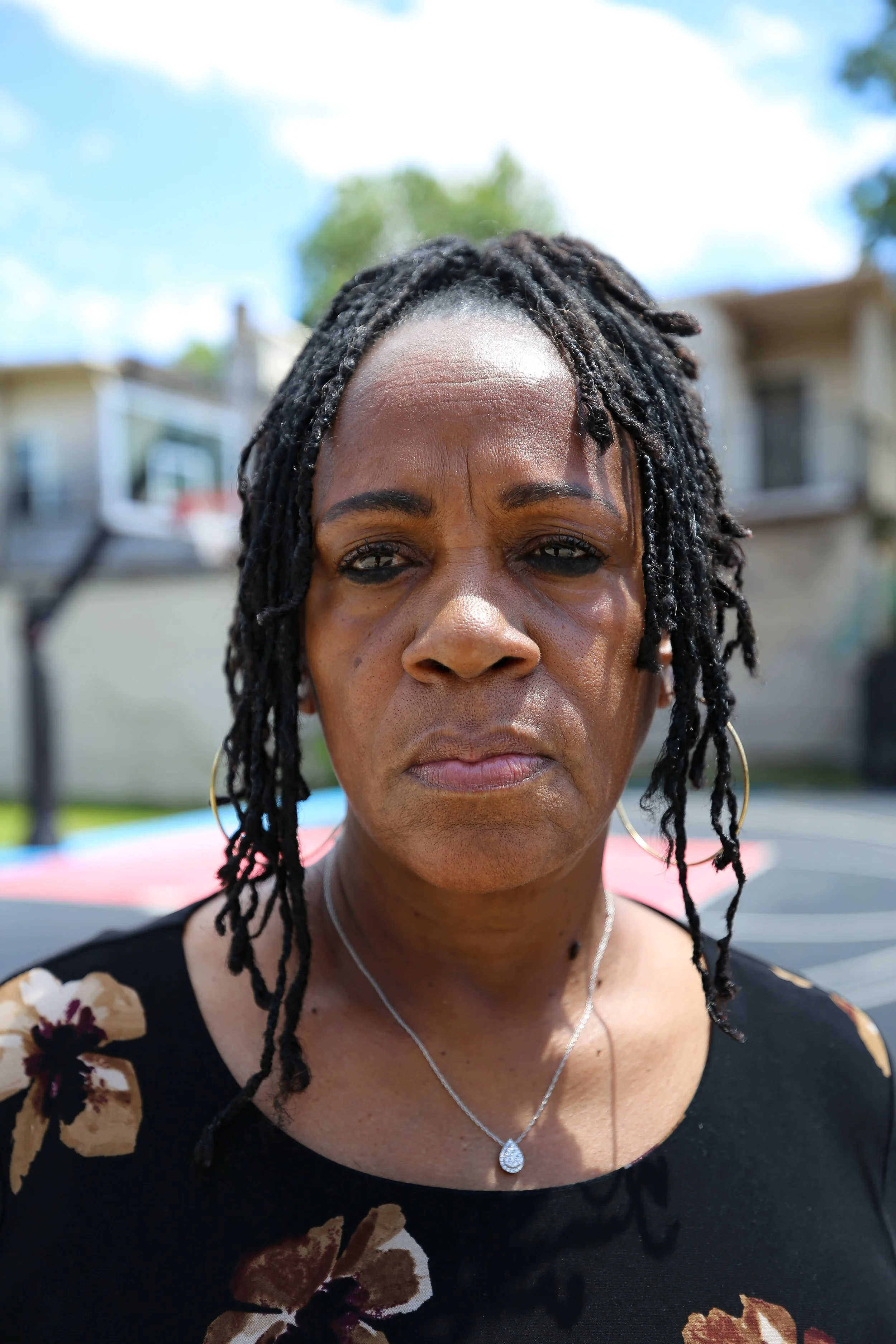 A woman with a serious expression, wearing a black floral shirt, hoop earrings, and a necklace, with dreadlocks and a basketball court in the background.