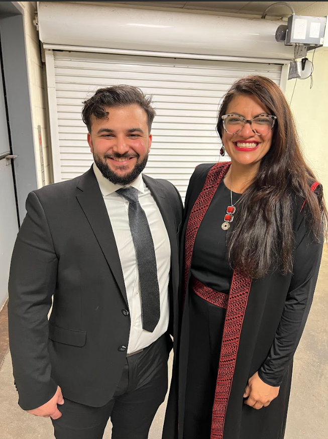 A man and woman dressed in formal attire standing together indoors in front of a closed roll-up door, smiling at the camera.