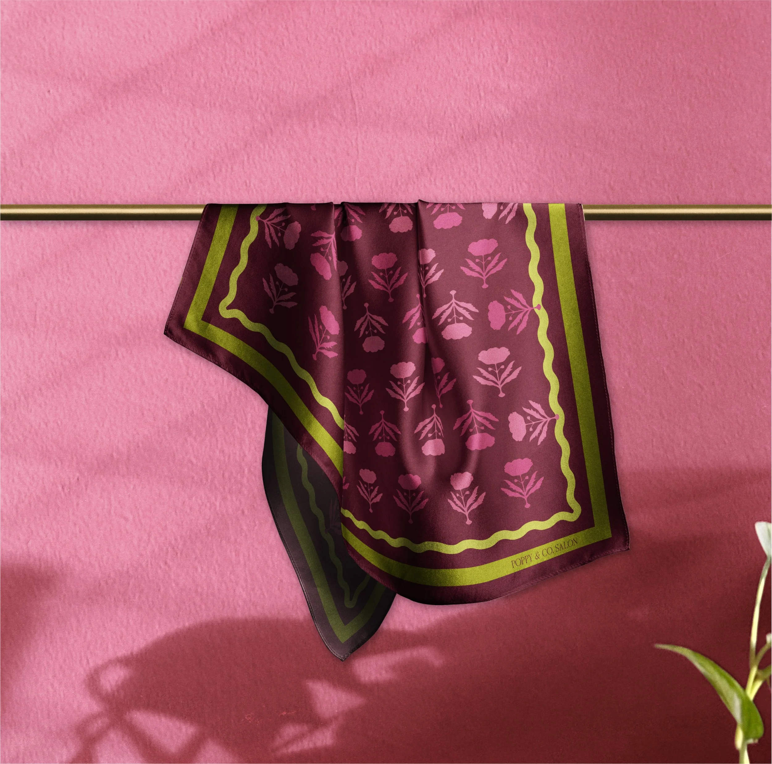 A maroon and green floral-patterned silk scarf hanging on a gold rod against a pink textured wall, with a small part of a green plant visible at the bottom right corner.
