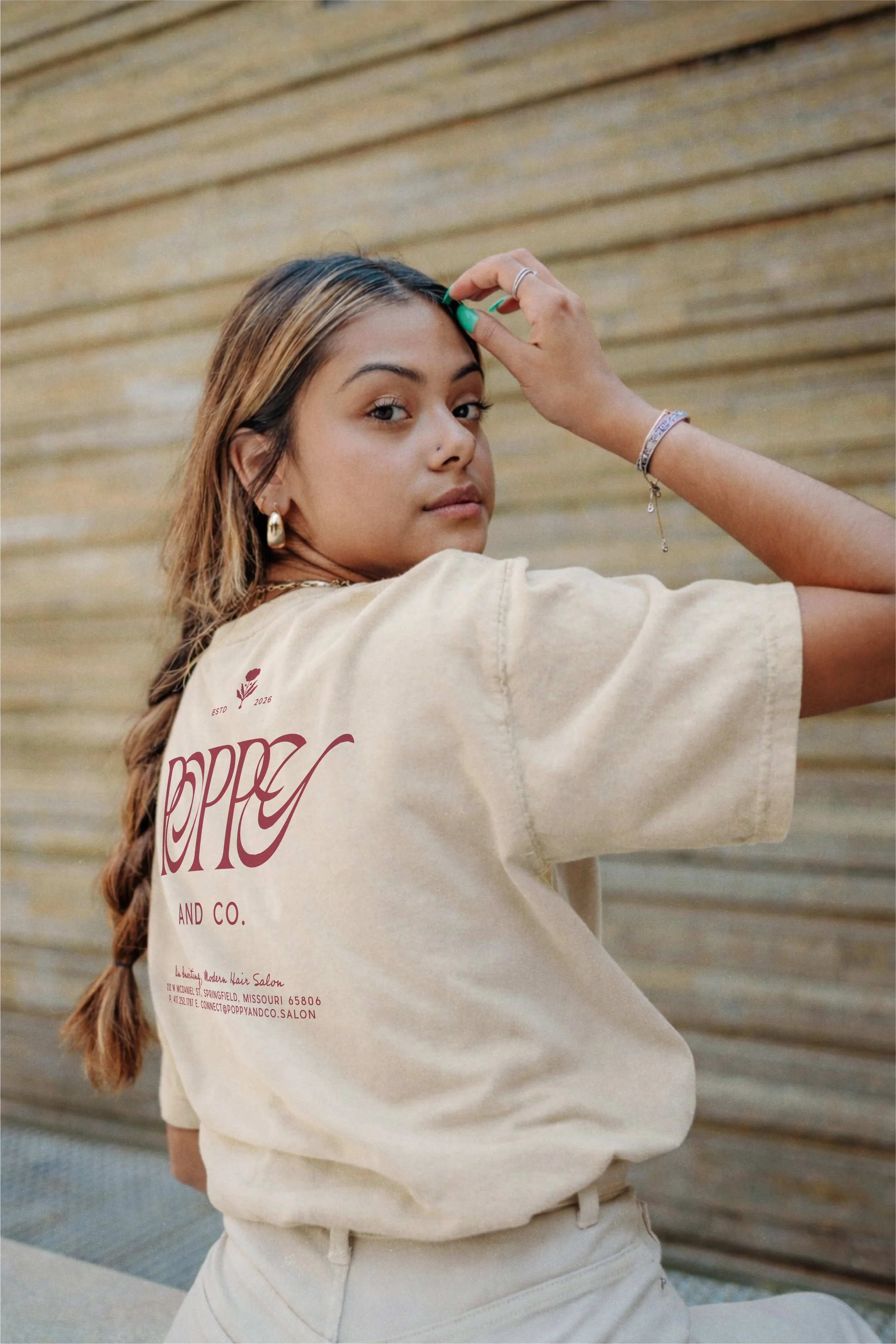 A young woman with light brown hair and hoop earrings looking over her shoulder, wearing a beige T-shirt with red text and standing against a wooden wall.