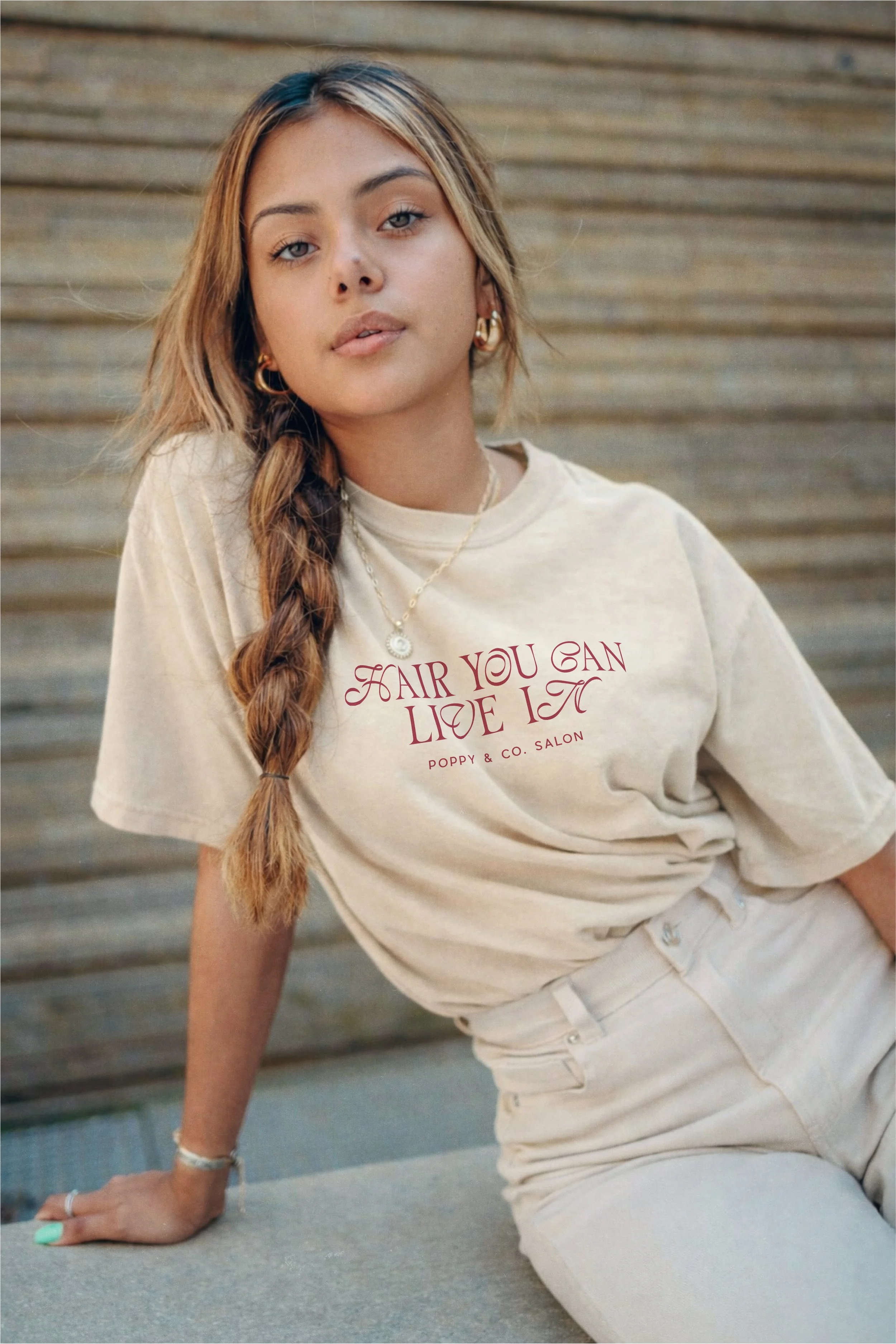 A young woman with long, wavy hair and gold jewelry posing outdoors against a wooden fence background. She is wearing a beige t-shirt with red text reading "Fair You Can Live Now" and light-colored pants.