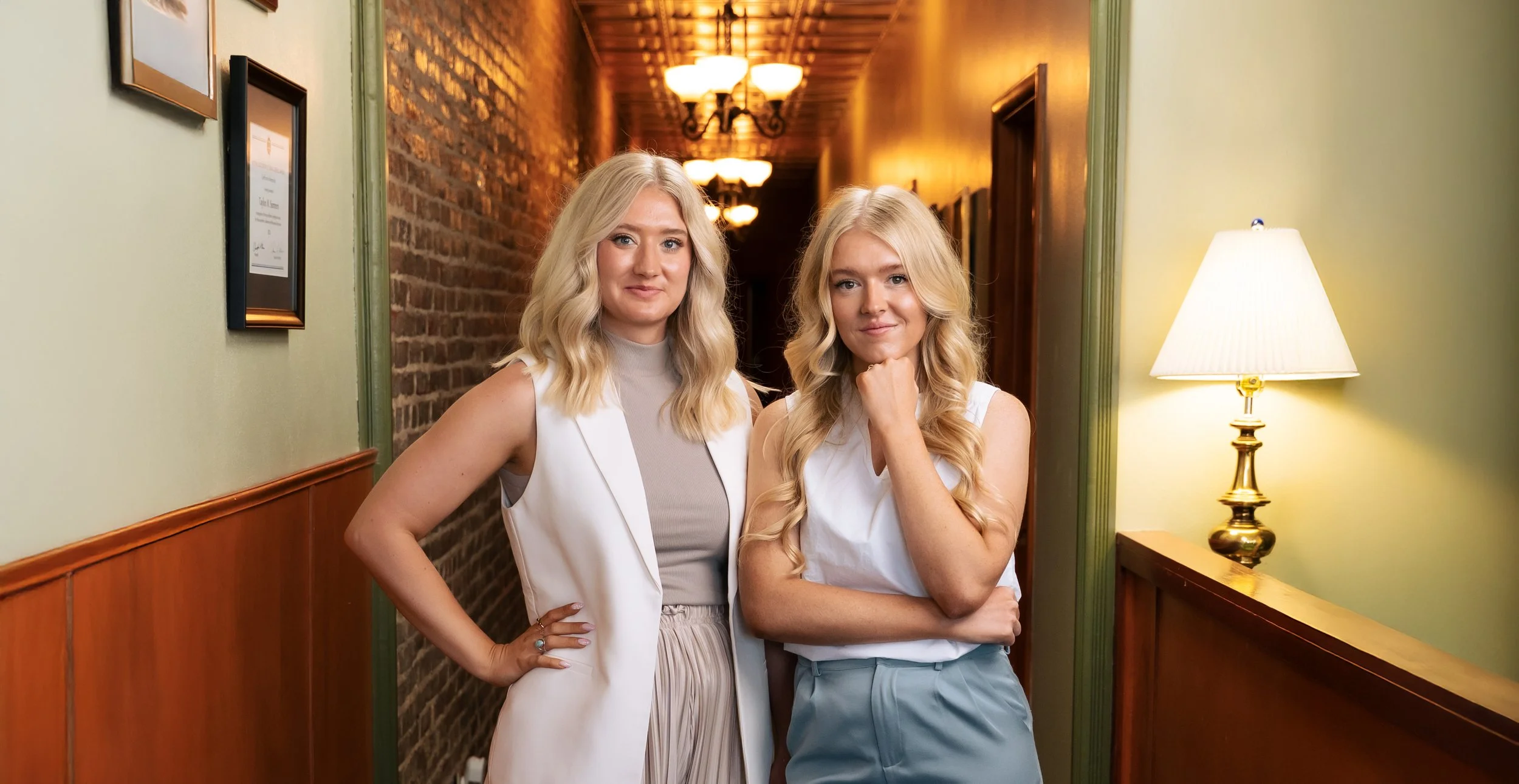 Two women with blonde hair and light skin standing in a hallway, one with a hand on her hip and the other with her hand on her chin, smiling at the camera. The hallway has brick and green painted walls, framed pictures, a wooden wainscot, and a lamp with a white shade.