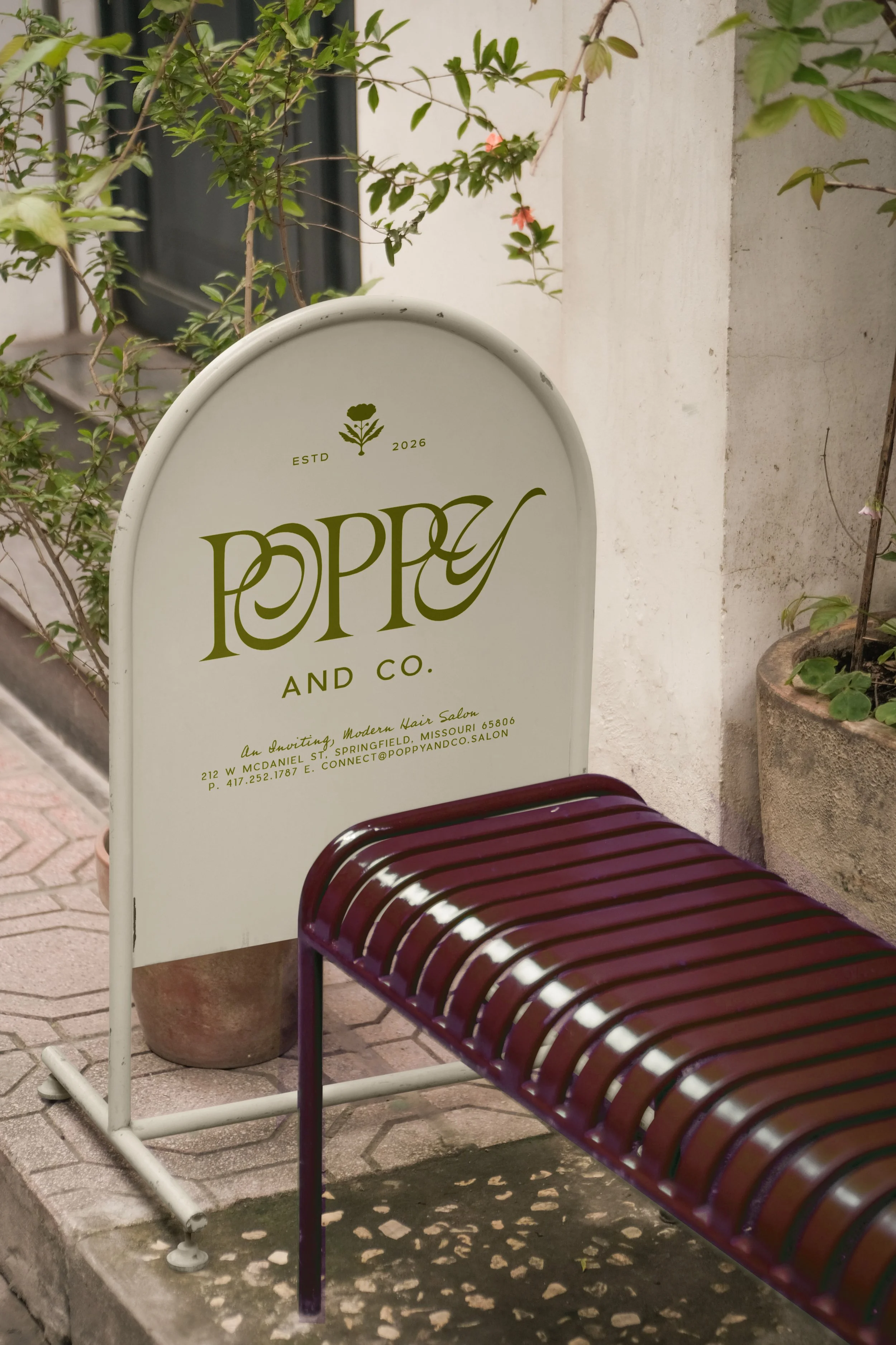 A white and burgundy outdoor sign for Poppy and Co., a modern hair salon, placed on a sidewalk next to potted plants.