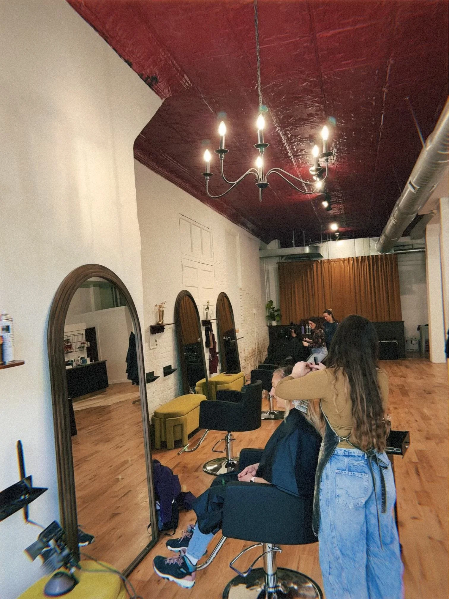 A hair salon interior with clients getting hair services, large mirrors, salon chairs, a chandelier, and a red ceiling.