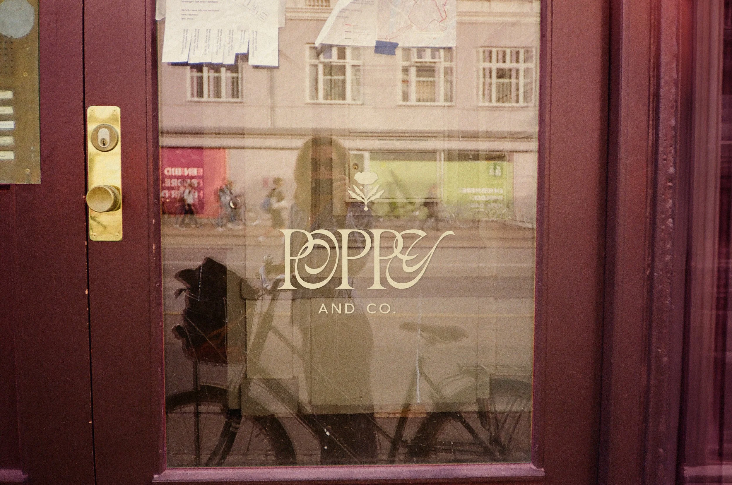 Storefront window with the logo 'Poppy and Co.' and a flower design, reflecting a bicycle, street scene, and people passing by outside.