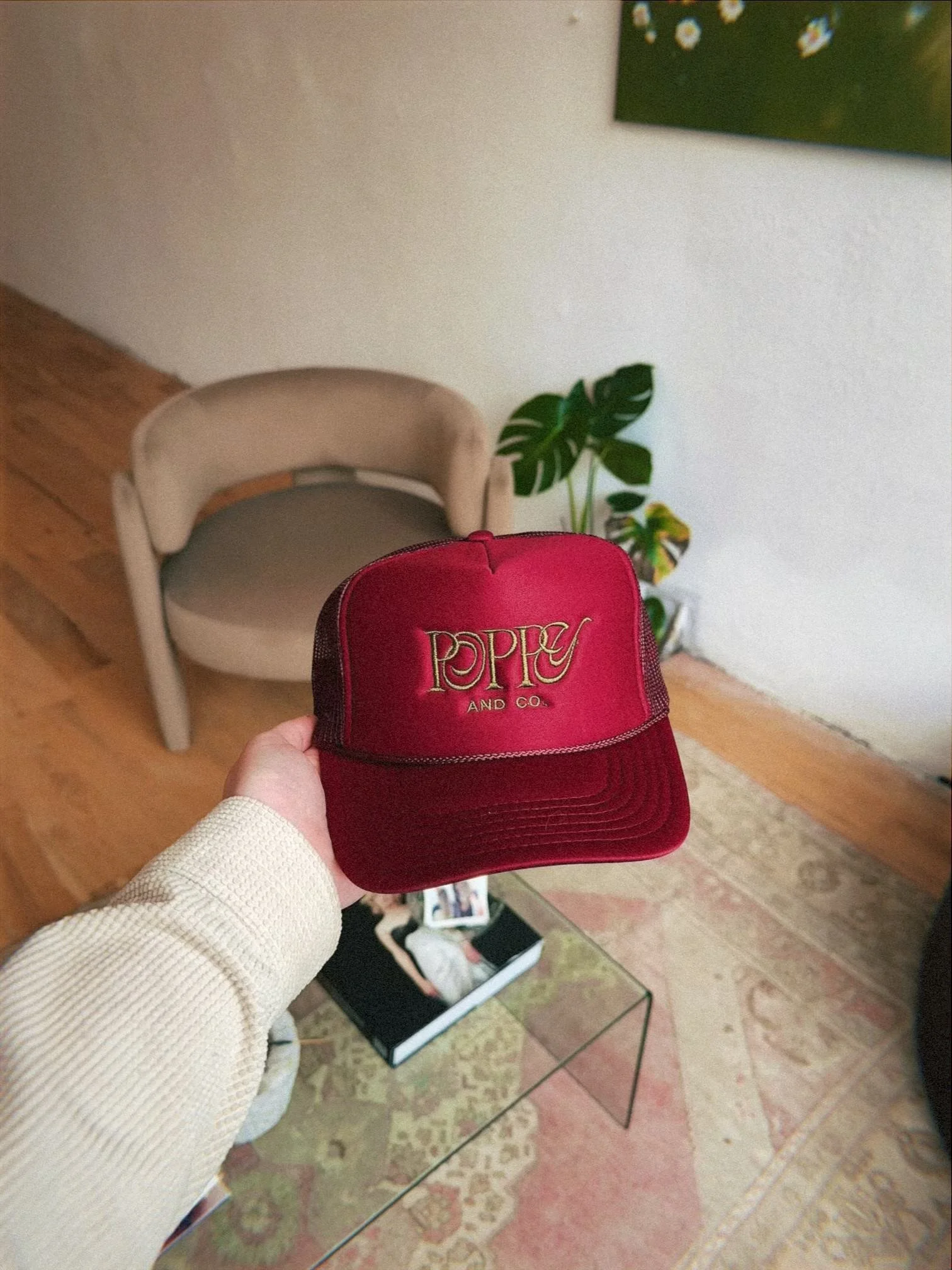 Hand holding a red trucker hat with gold embroidery that says 'POPPA AND CO.' in a living room with a beige chair, a potted plant, a glass table, and a large wall art piece.