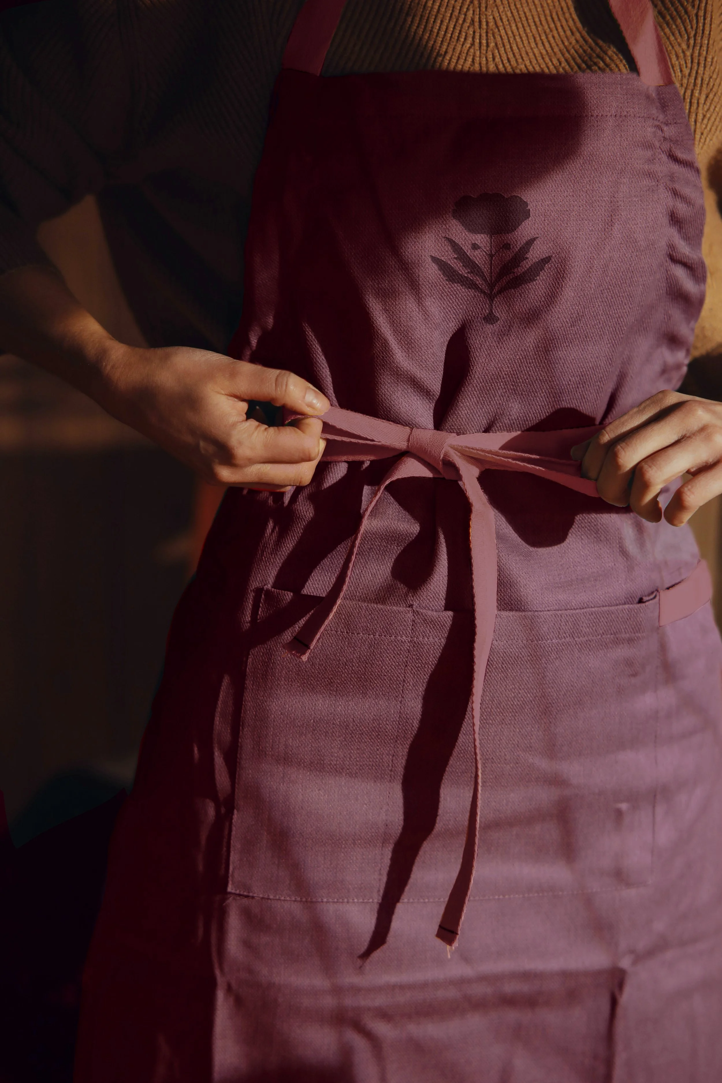 A person tying the waist of a pink apron with a dark flower design on it.