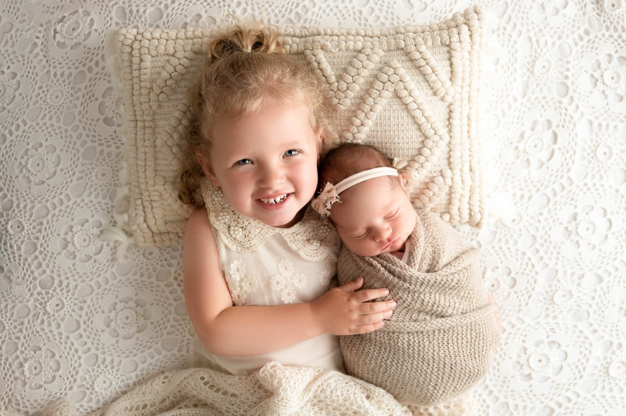 toddler laying on back hugging newborn sister resting heads on cream pillow