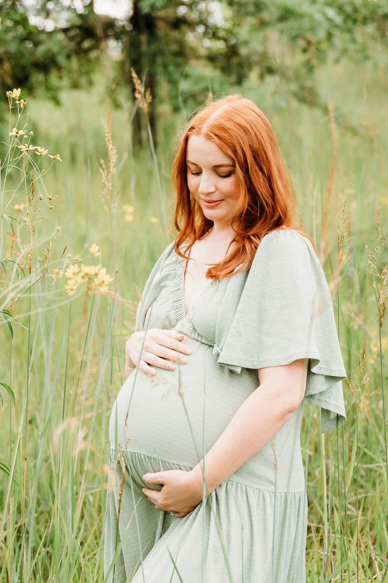 Pregnant red-haired woman in a sage green dress holding her belly in a grassy field during an outdoor maternity session in Indianapolis