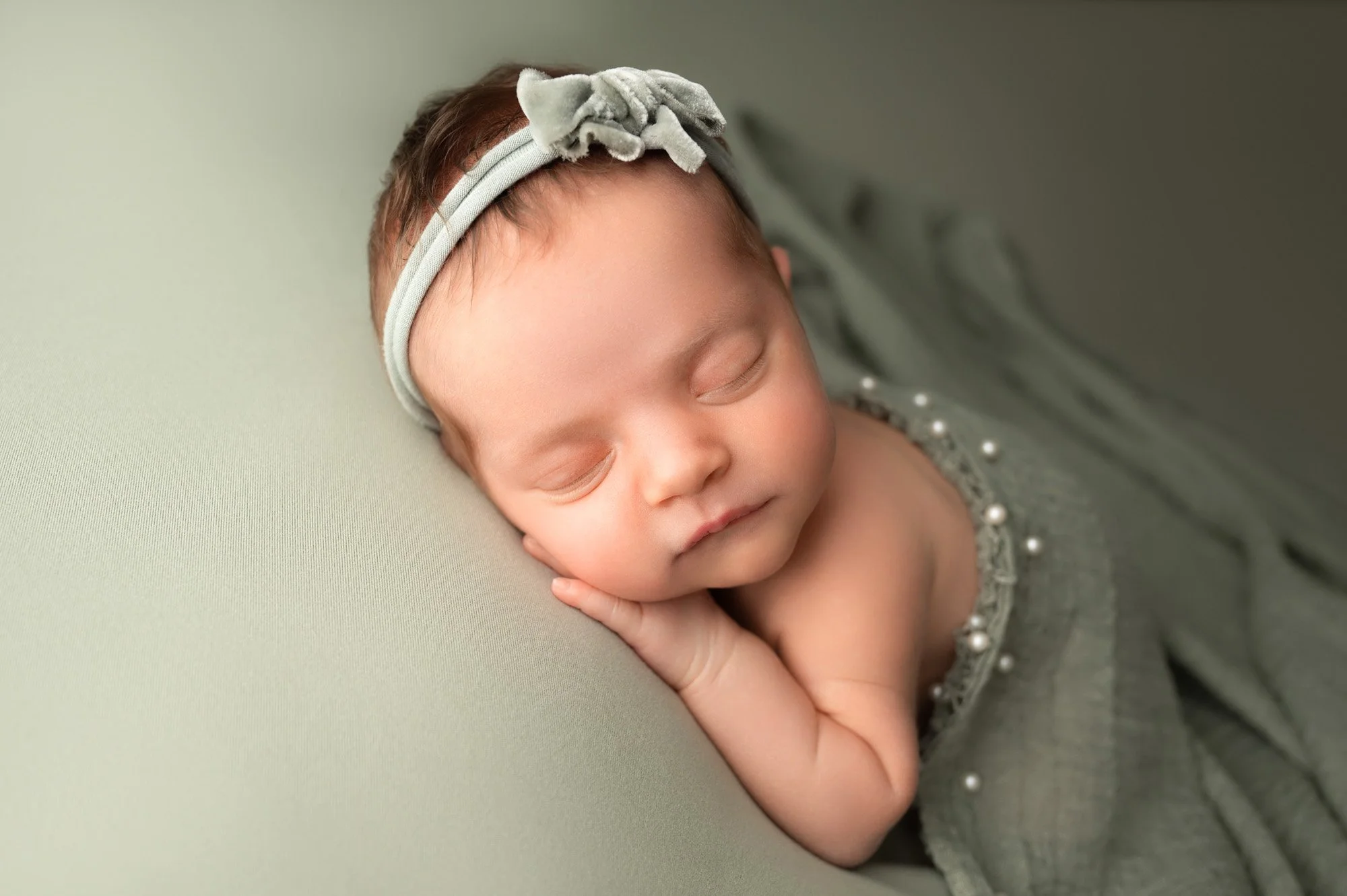 Sleeping newborn posed on sage green backdrop with hands tucked under cheek and matching velvet bow headband in Indianapolis studio
