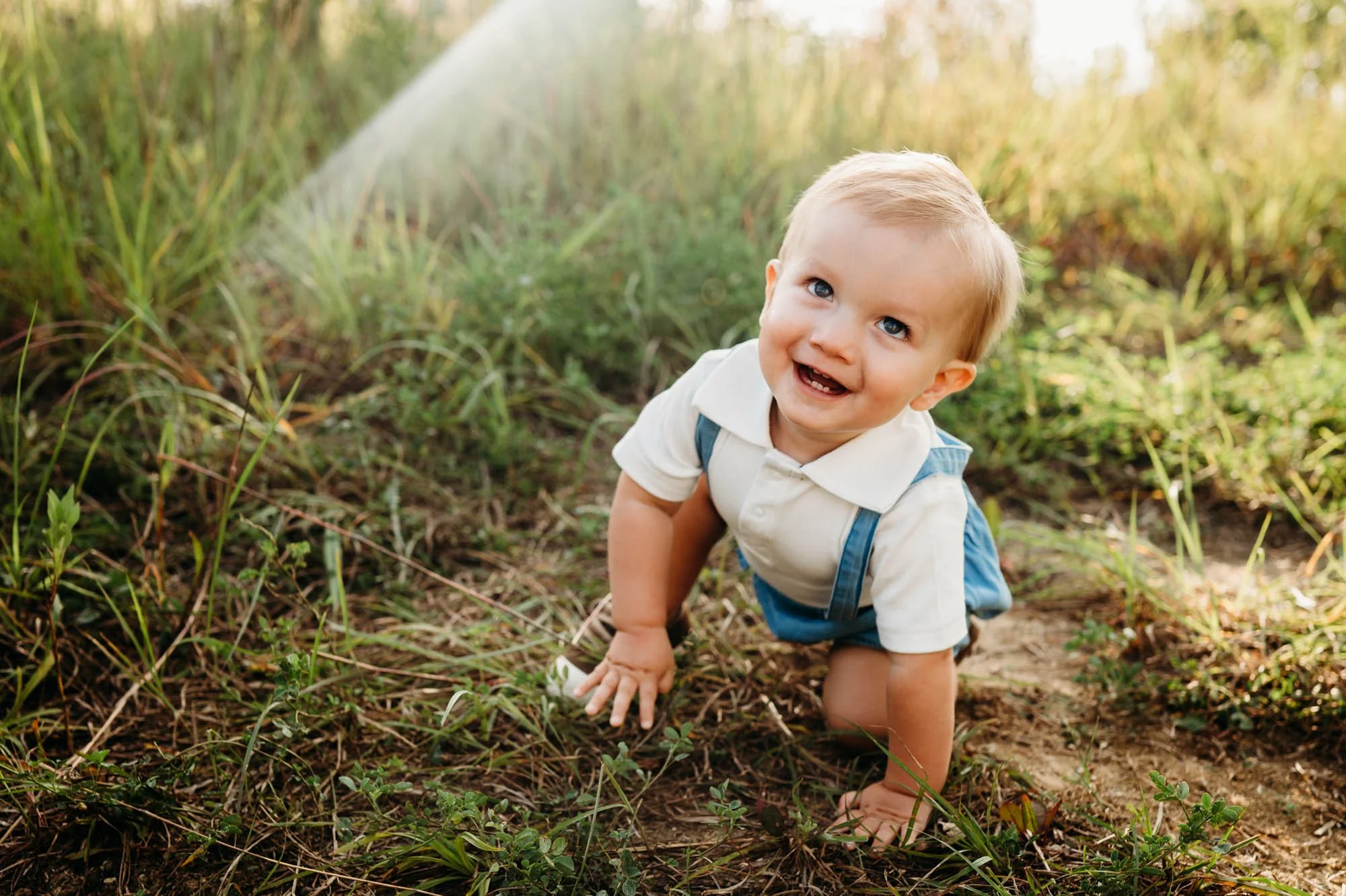 Smiling baby in blue suspenders crawling on the ground during an outdoor family session in Noblesville, Indiana