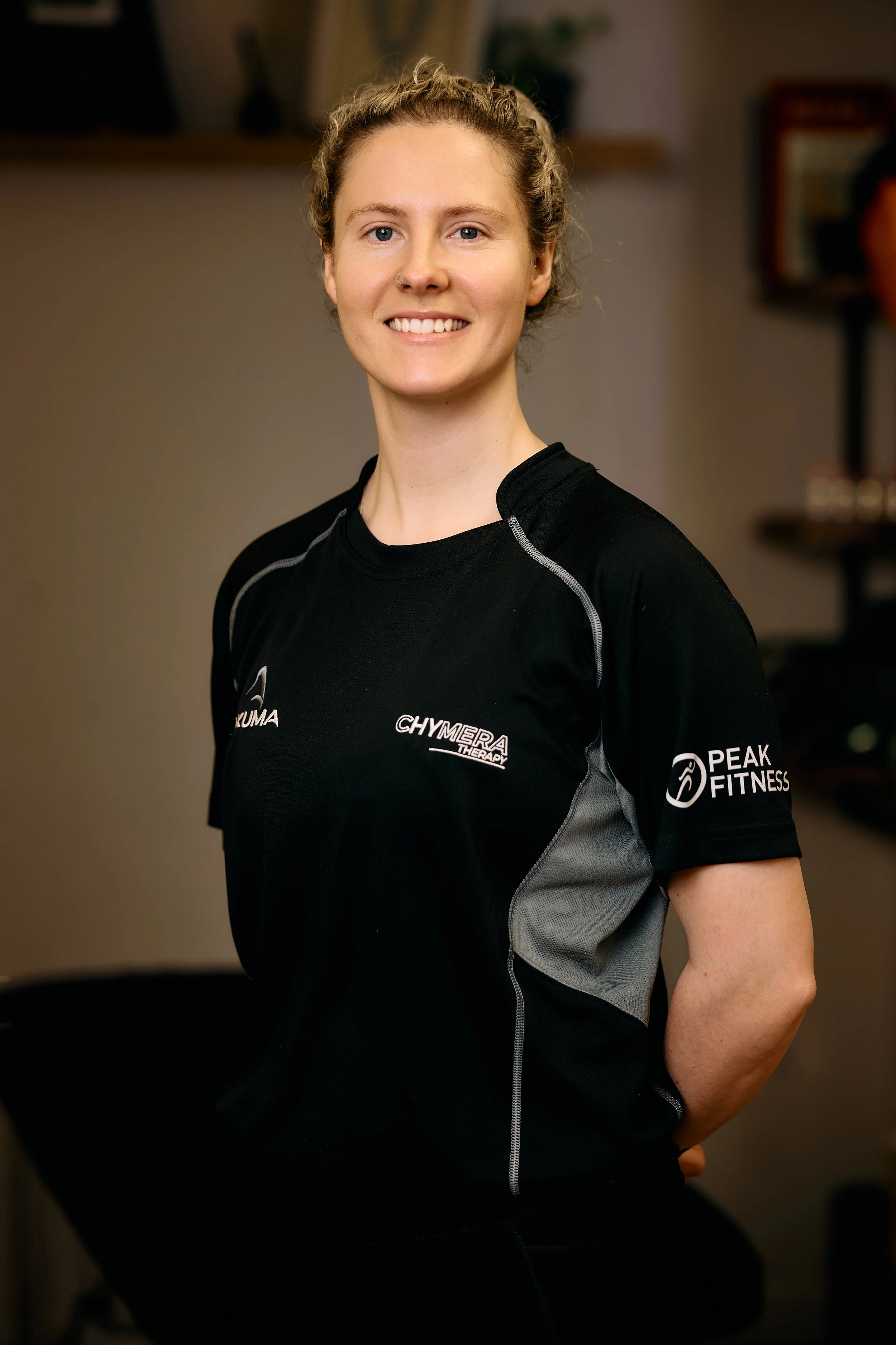 Jess, the owner of Chymera Therapy, a smiling woman with blonde curly hair tied back, wearing a black athletic shirt with logos, standing indoors.