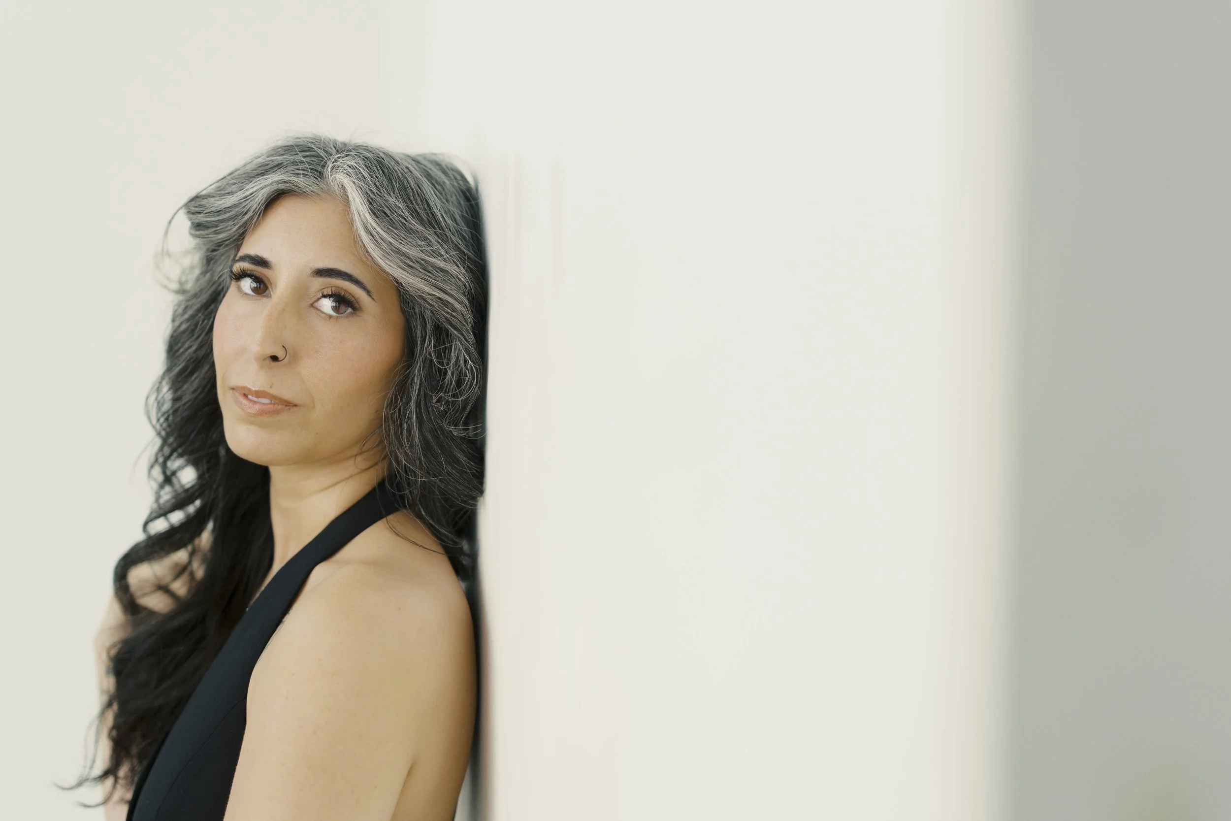 A woman with long dark hair, gray streaks, and a nose piercing, wearing a black top, looking at the camera against a plain light-colored background.