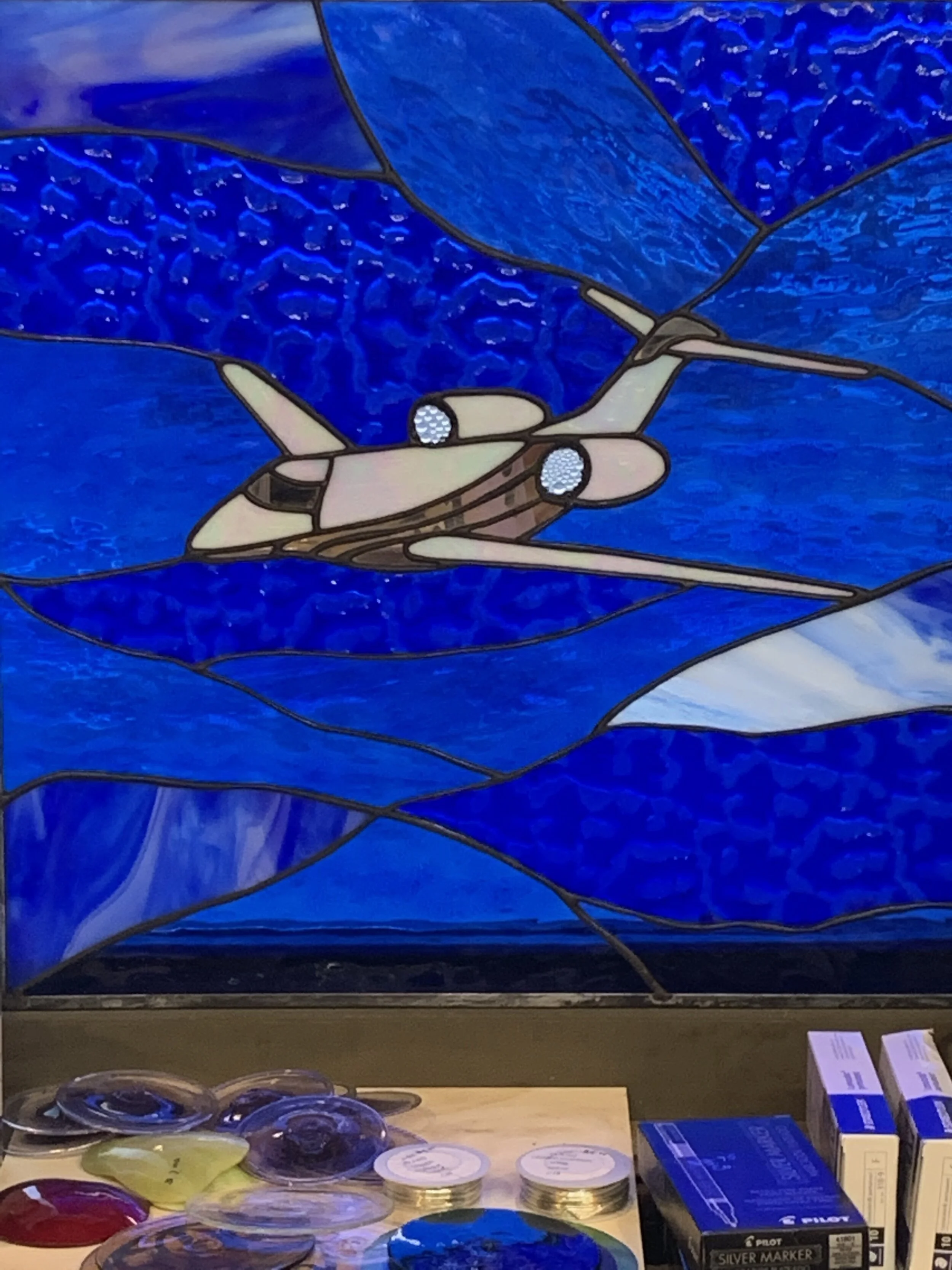 Colorful custom designed stained glass artwork depicting white plane jet flying over blue sky white clouds 