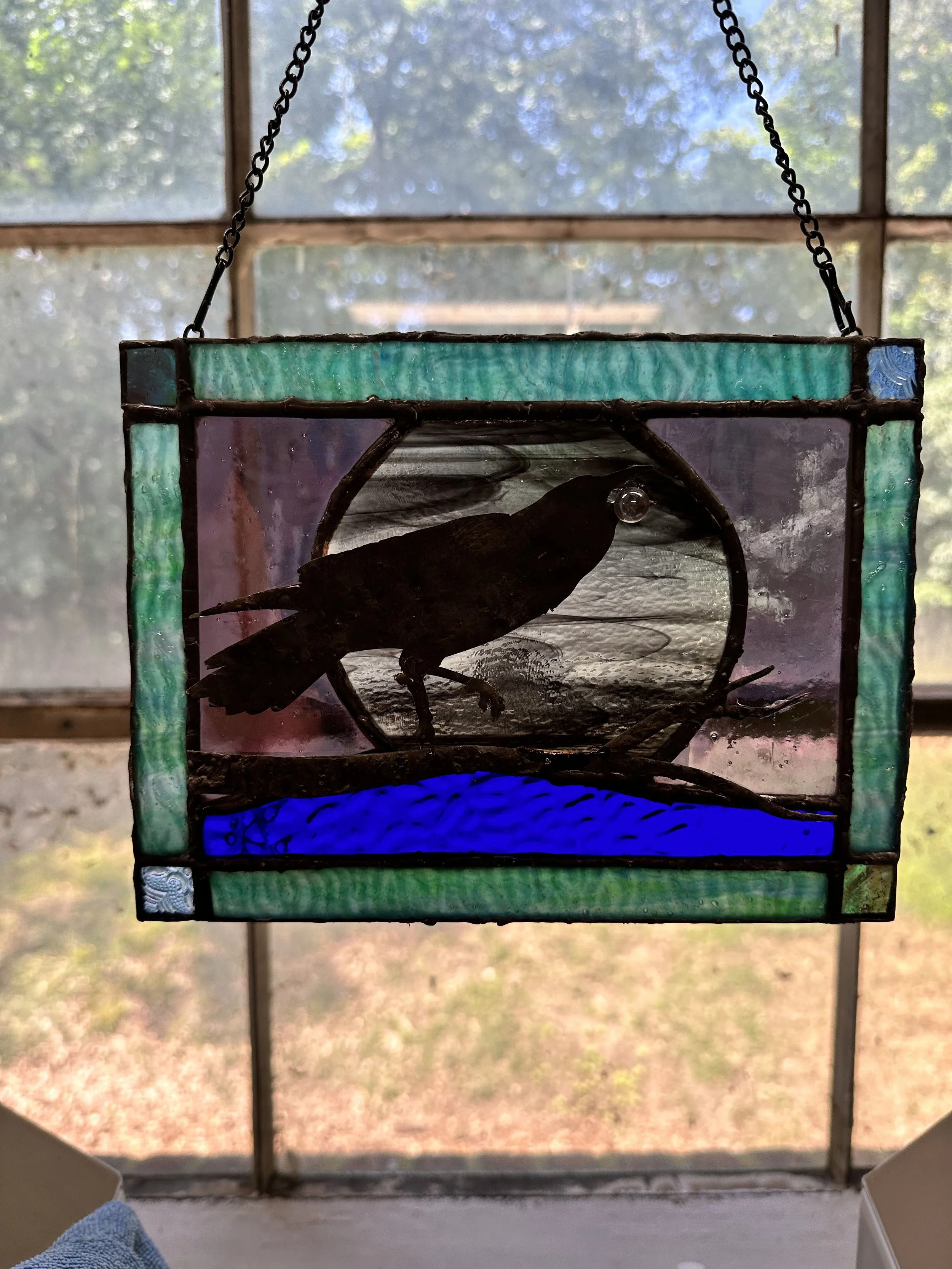 Stained glass hanging ornament featuring a black silhouette of a bird, possibly a raven or crow, perched on a branch above blue water, with a background of purple and green glass.