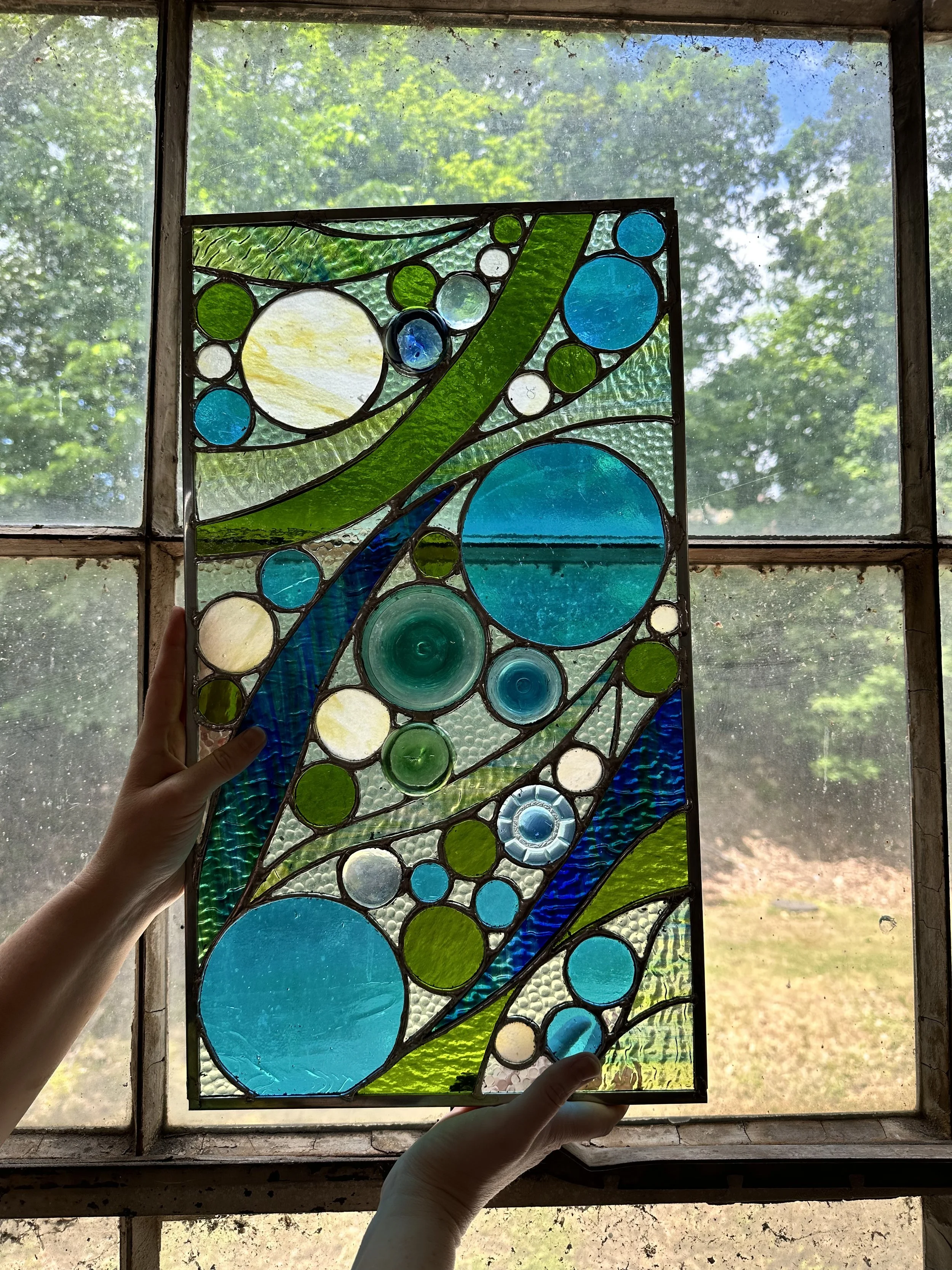 Person holding a stained glass window panel with blue, green, white, and clear glass circles and curved shapes in front of a window showing trees and sky outside.