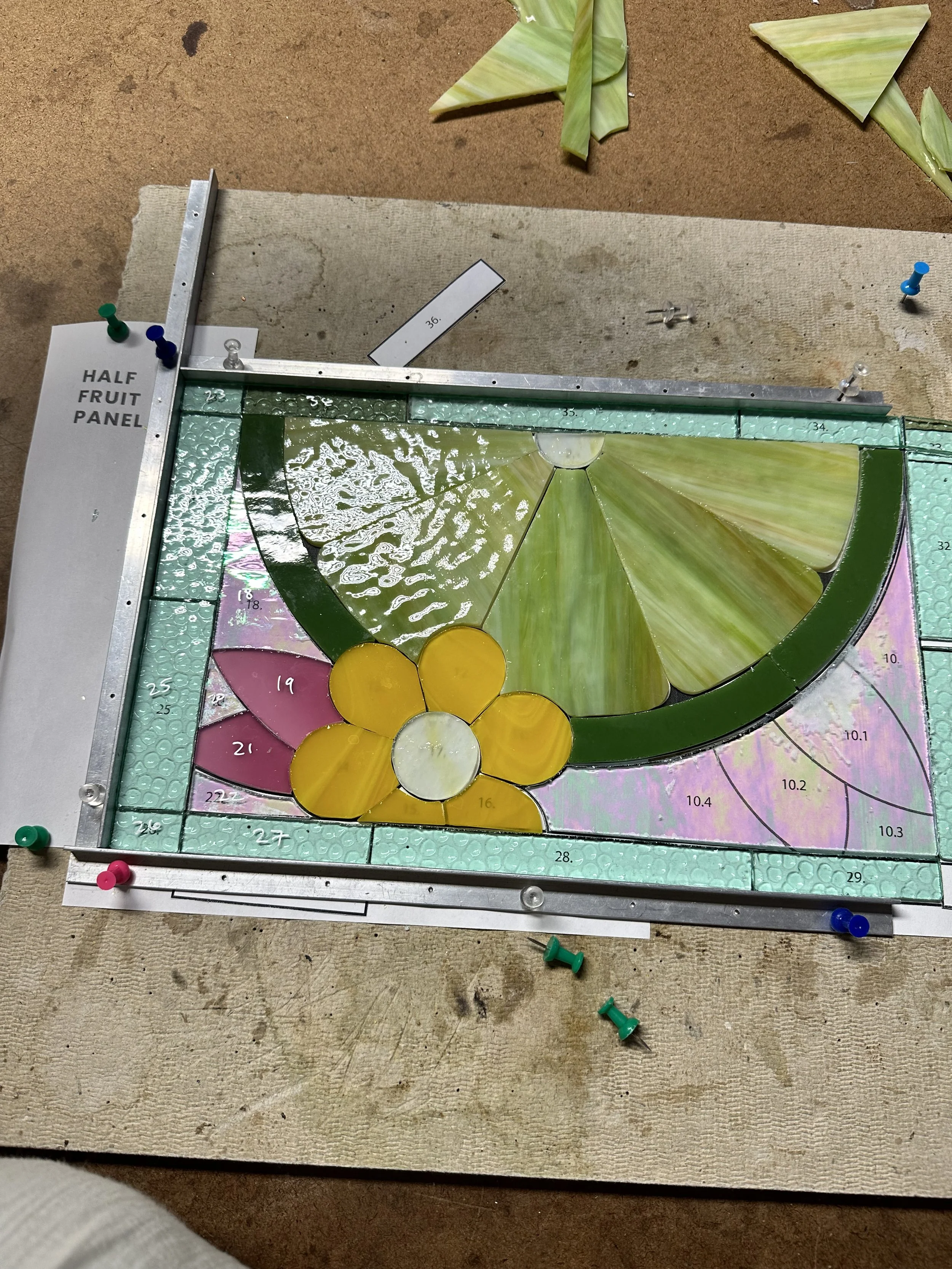 A stained glass panel in progress depicting a yellow flower with pink petals and a green stem, labeled as "Half Fruit Panel" with various measurement markings.