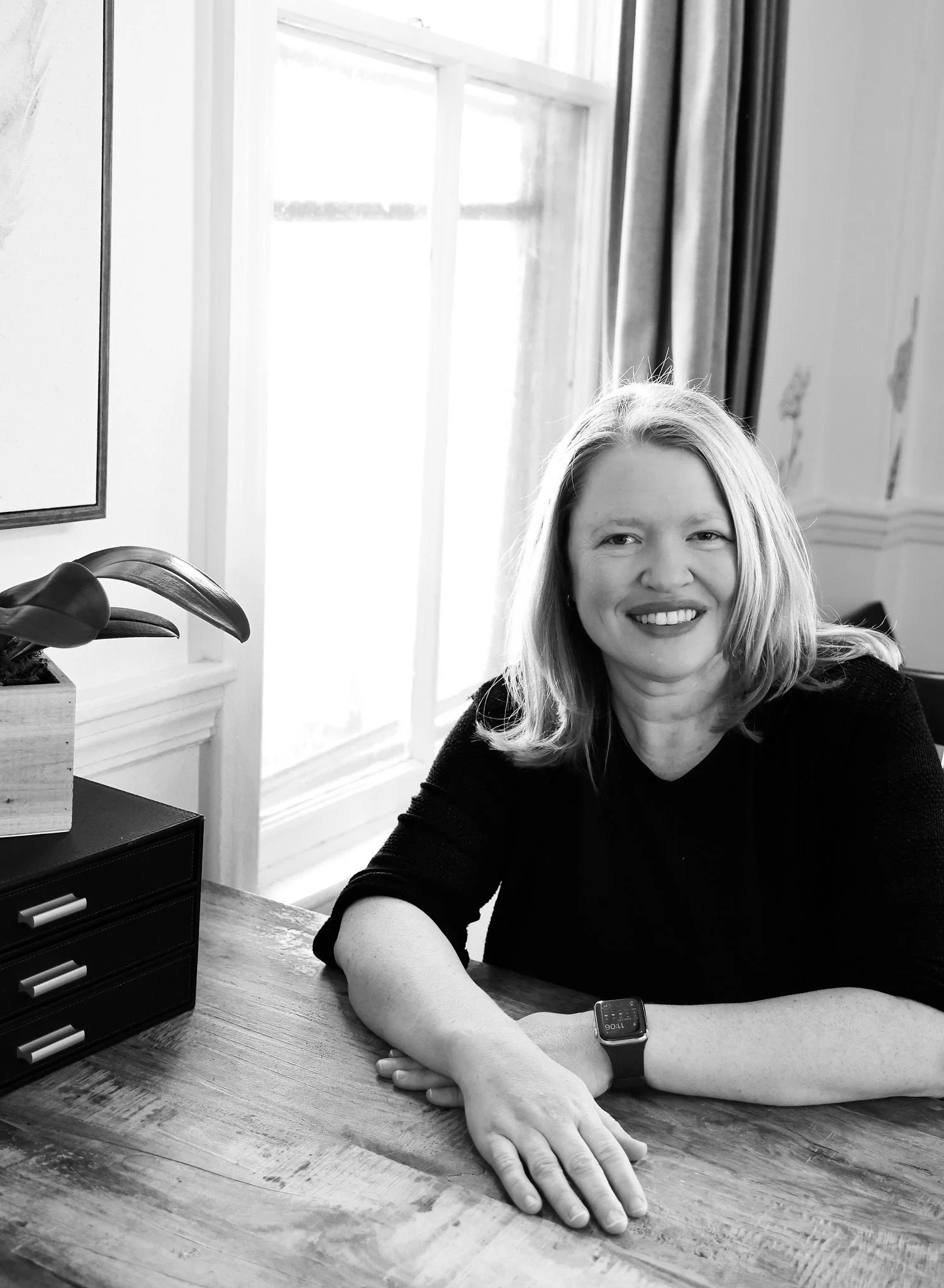 Black and white portrait of a woman with blonde hair, smiling, sitting at a wooden table next to a window, with a small plant in a square pot and a black storage box nearby.
