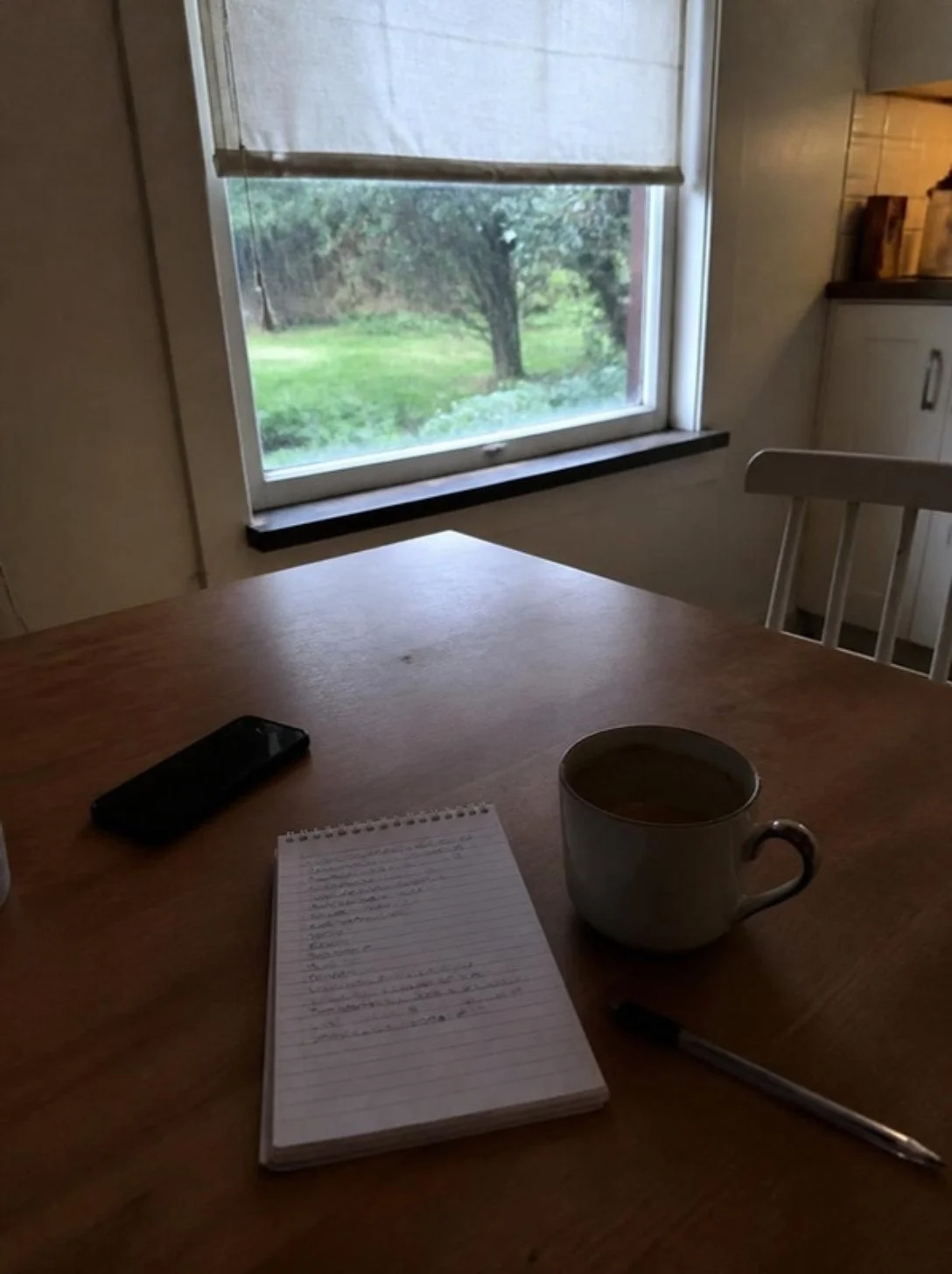 Anteckningsblock, mobil och kaffekopp på ett köksbord vid fönstret