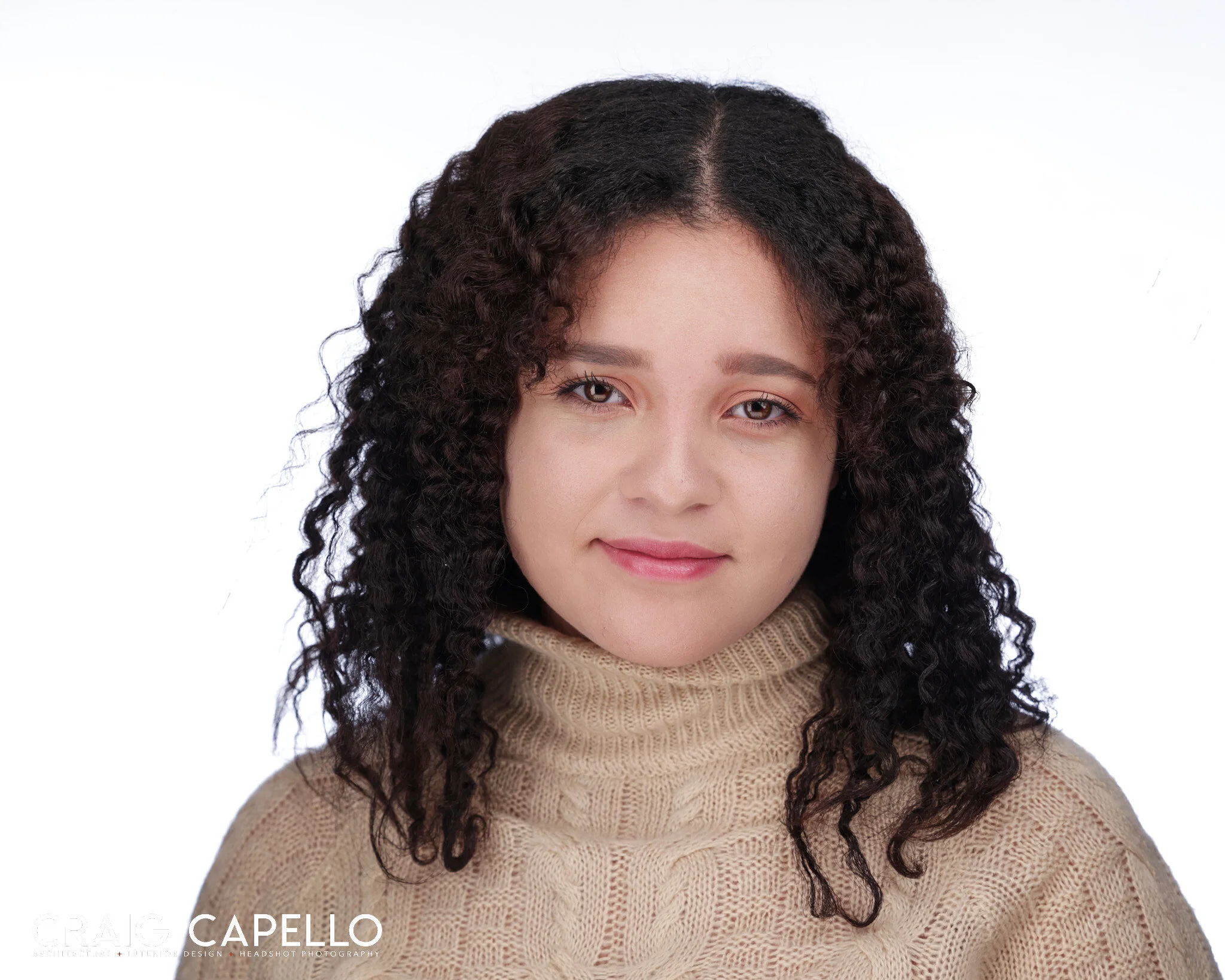 Smiling person with curly hair wearing a beige sweater against a white background.