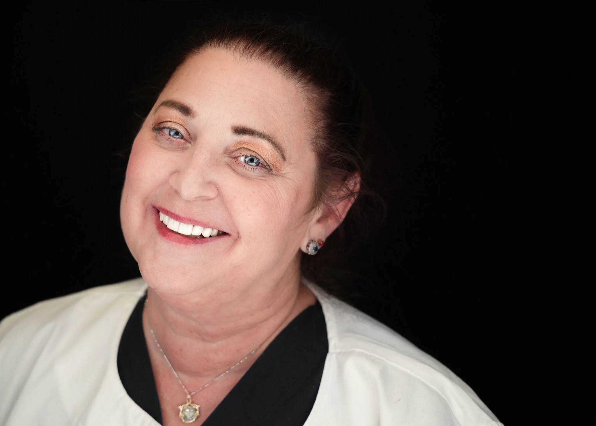 Smiling woman with dark hair wearing a necklace and a light-colored top against a black background.