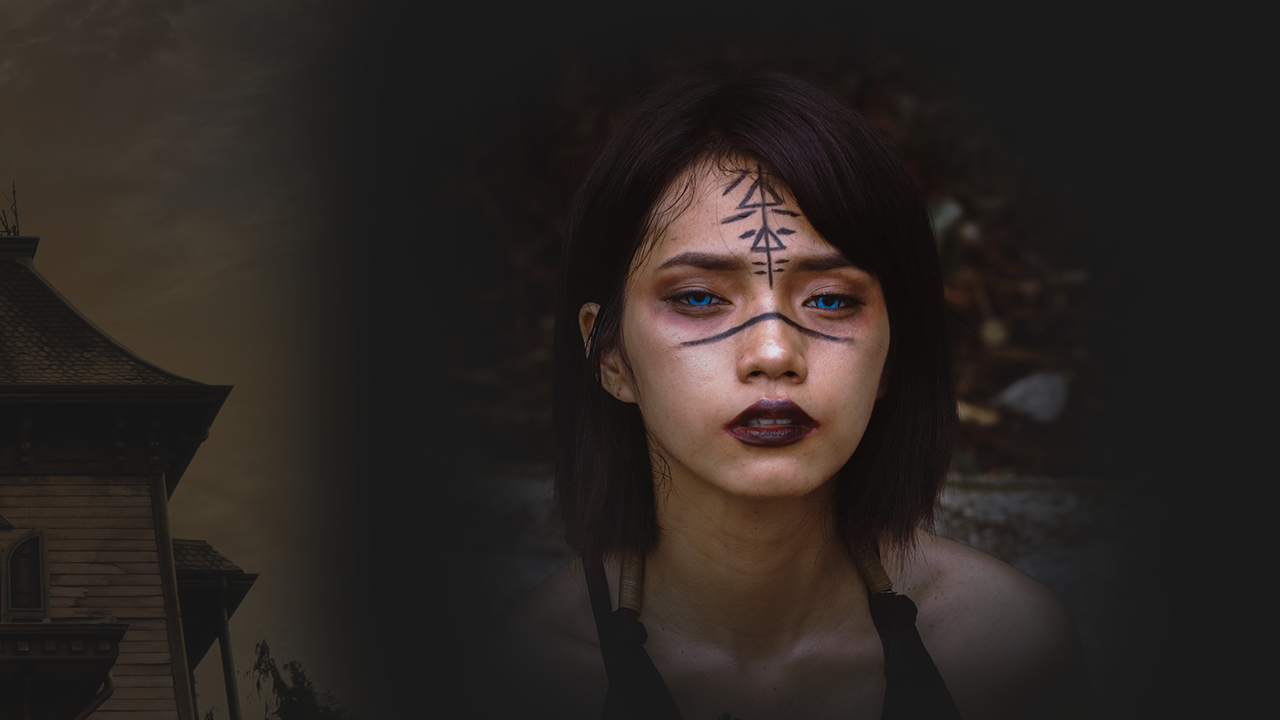 Woman with tribal-style face paint, dark makeup, and dark hair, outdoors during daytime, with a house silhouette partially visible on the left.