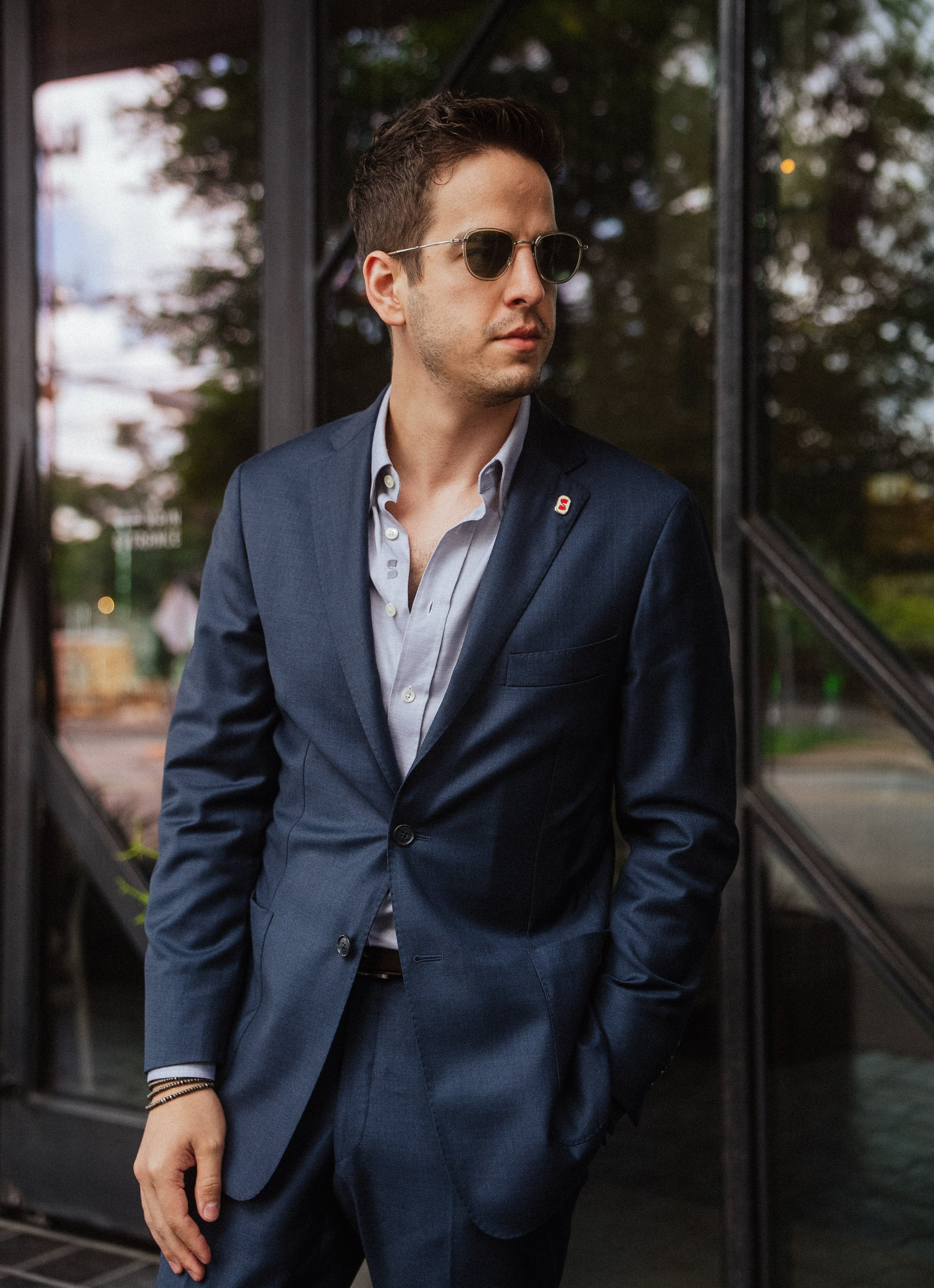 Man in a navy suit and sunglasses standing outdoors, looking to the side.