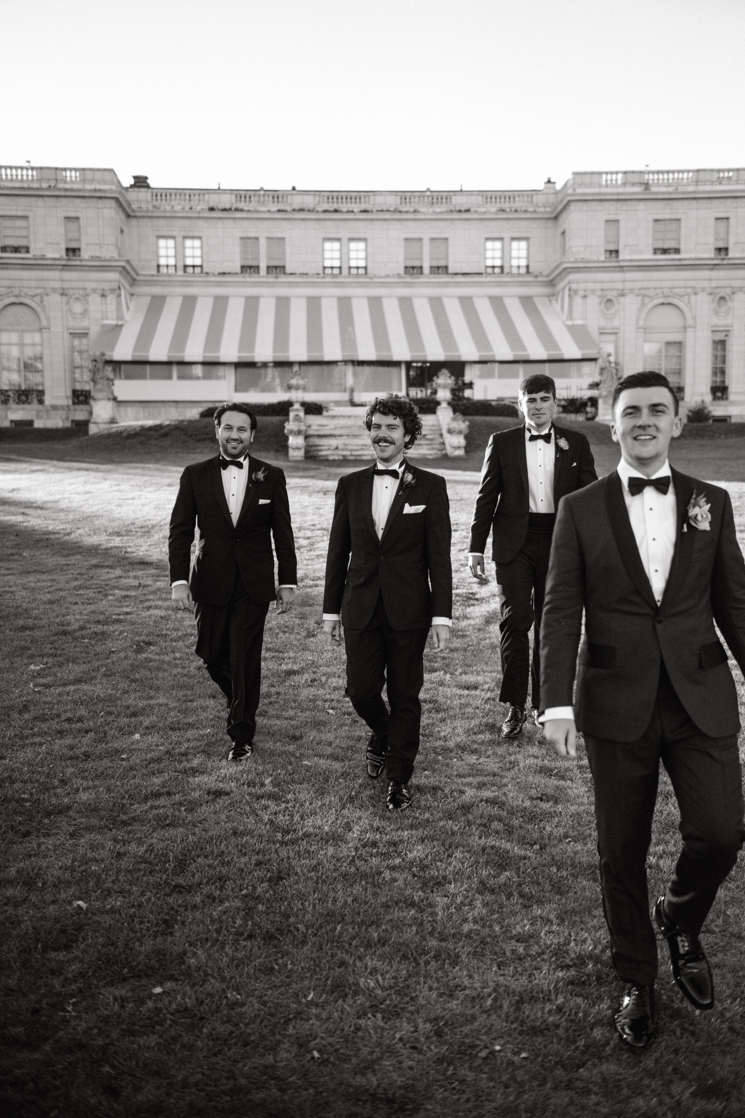Four men in tuxedos walking on grass outside a large building with striped awning, black and white photo.