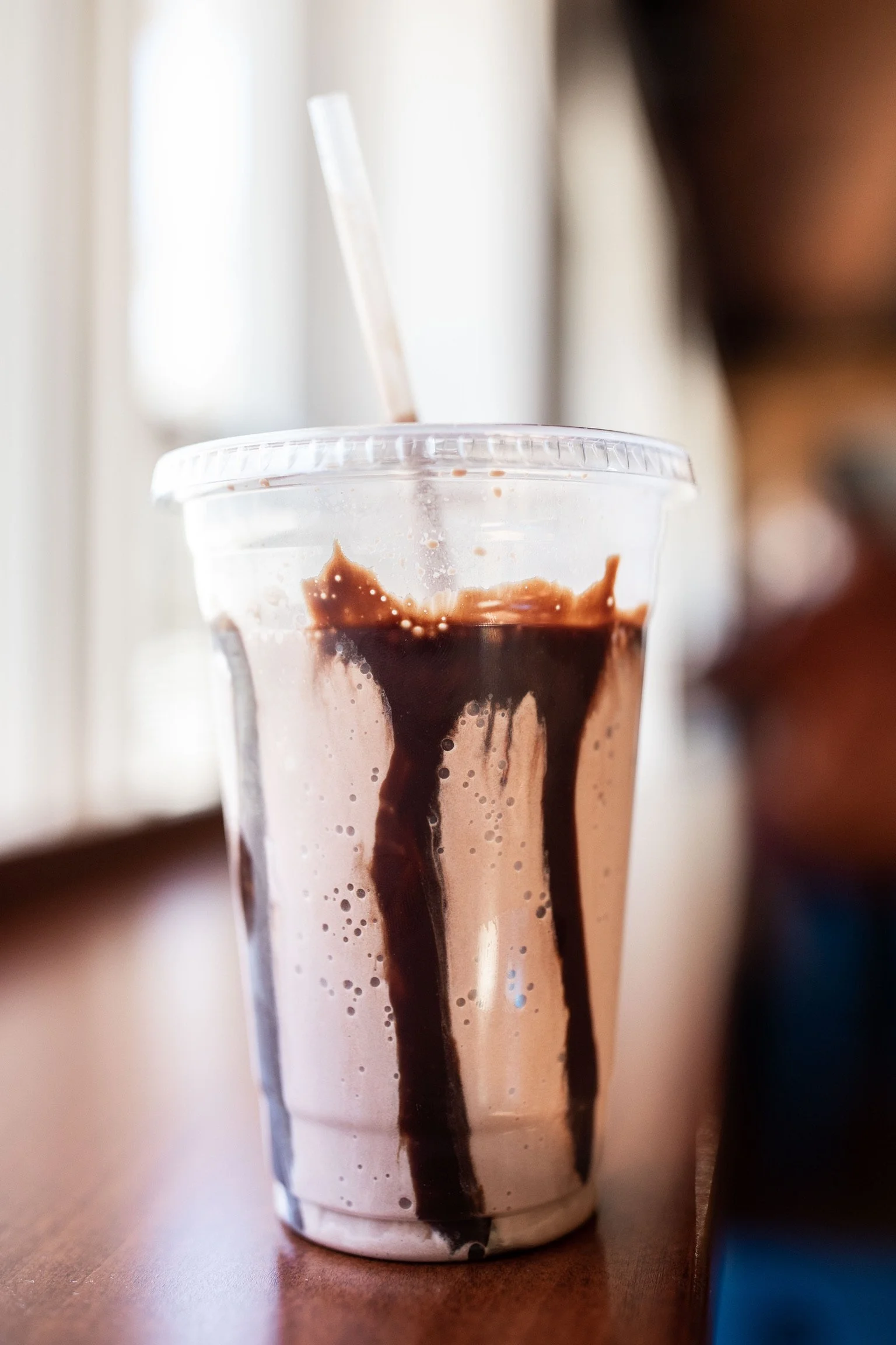 Close-up of a plastic cup filled with dark iced chocolate with chocolate syrup dripping down the sides, placed on a wooden surface.