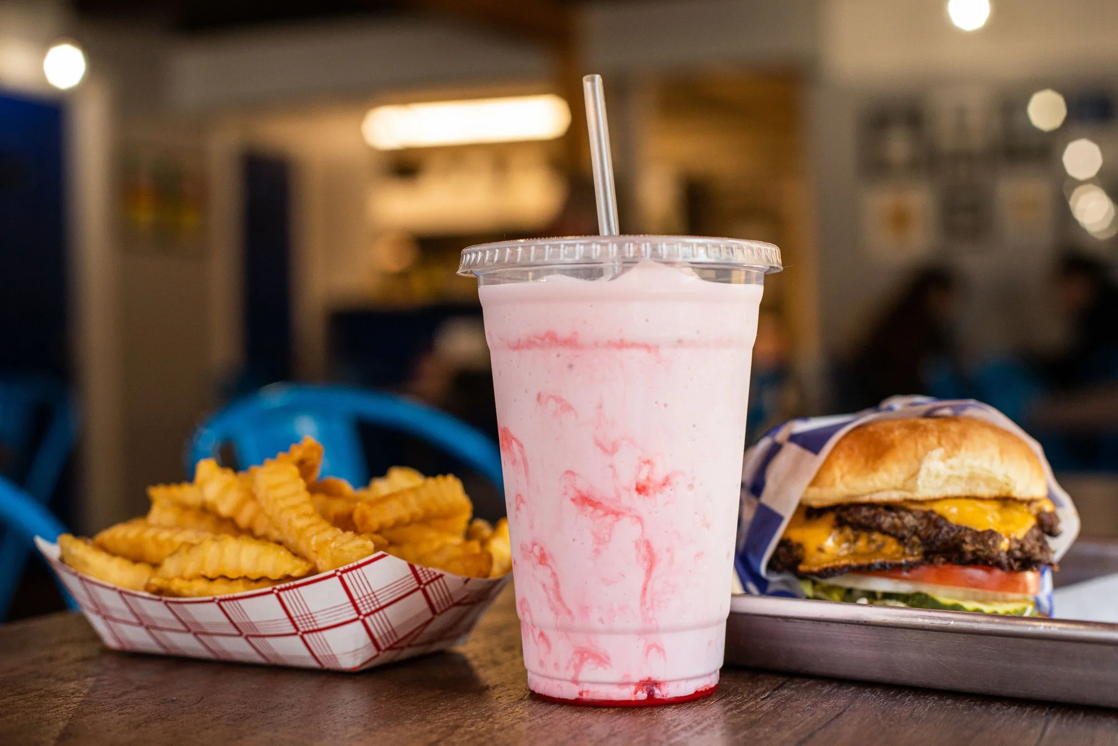 A pink and white milkshake with a straw in a plastic cup, served with crinkle-cut fries in a paper tray, and a cheeseburger with lettuce, tomato, and cheese on a toasted bun, in a casual restaurant setting.