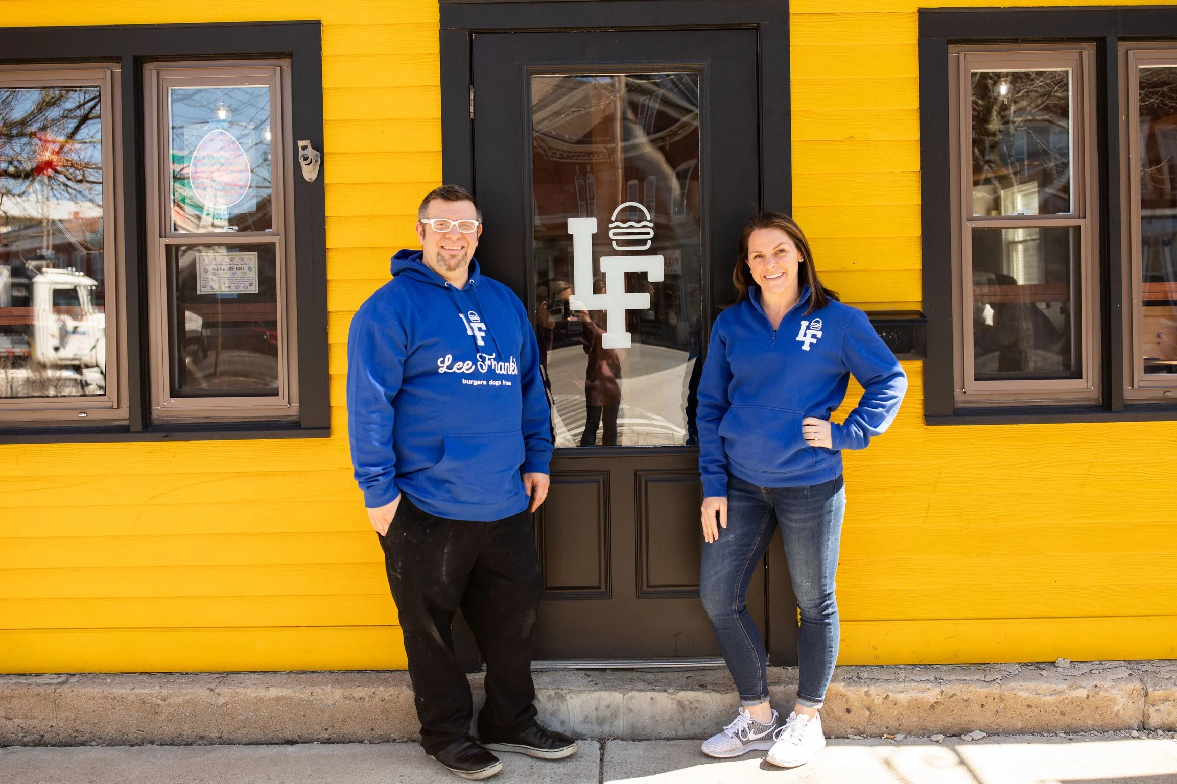 Two smiling people, a man and a woman, standing outside in front of a yellow building with black trim and large windows, wearing blue hoodies with white text.