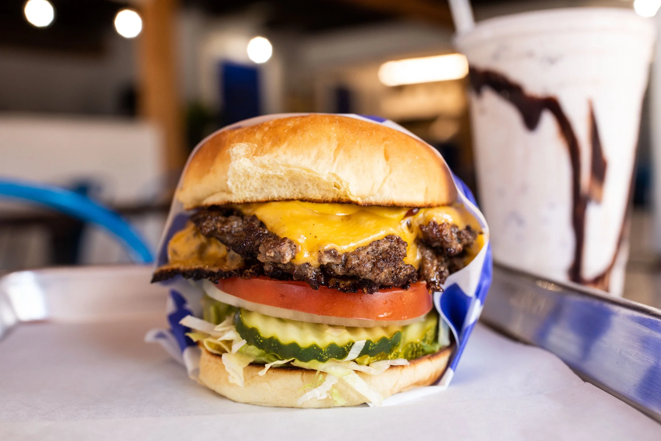 A cheeseburger with lettuce, tomato, pickles, onion, and two beef patties on a bun, with a cup of iced beverage in the background.