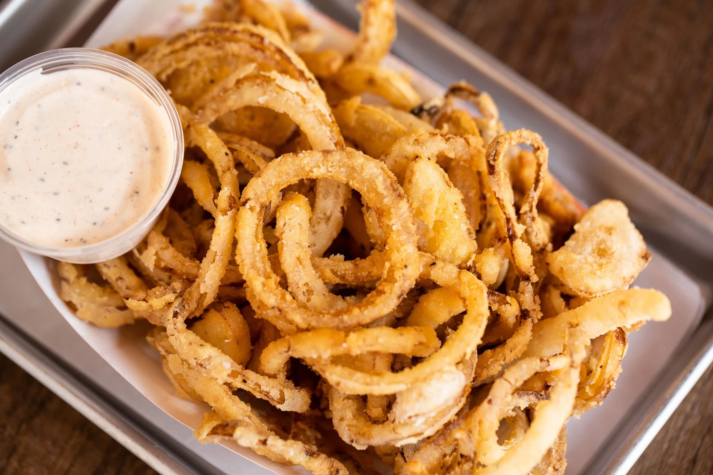 A serving of crispy fried onion rings with a side of creamy dipping sauce in a small cup, placed on a tray.