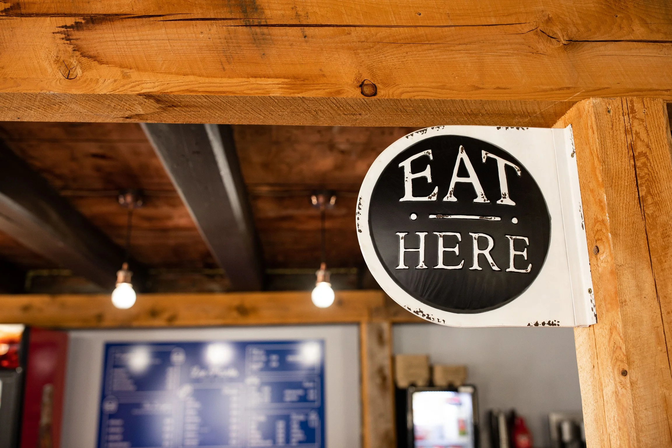 Wooden sign with black and white text that reads 'EAT HERE' mounted on a rustic wooden beam.