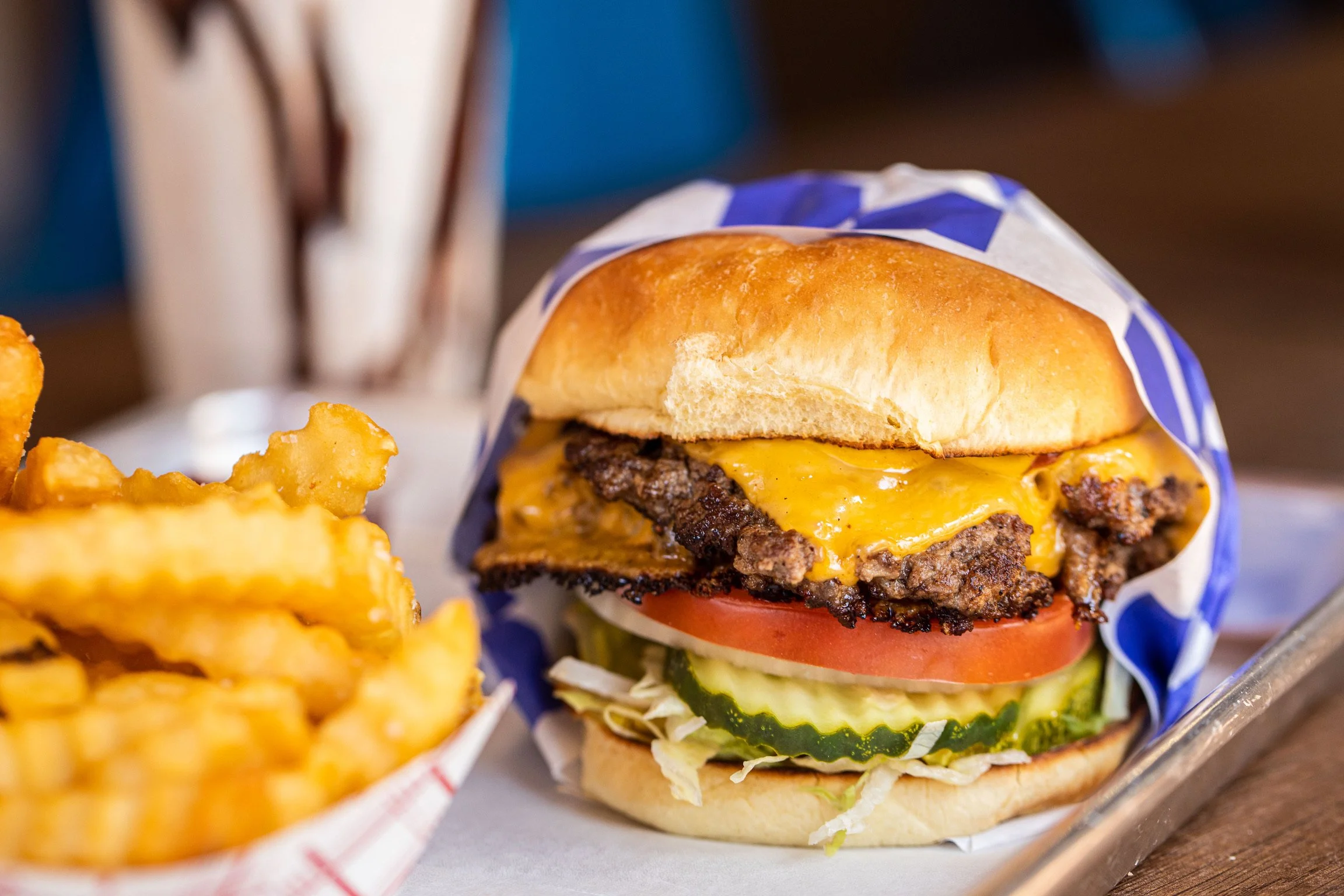 Close-up of a cheeseburger with lettuce, tomato, pickles, onions, melted cheddar cheese, and a beef patty in a bun, served with crinkle-cut fries on a tray.