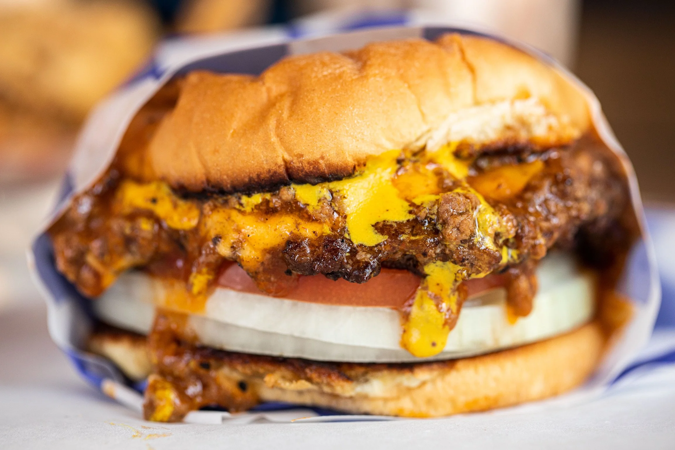 Close-up of a cheeseburger with a toasted bun, melted cheese, beef patty, and toppings, wrapped in paper.
