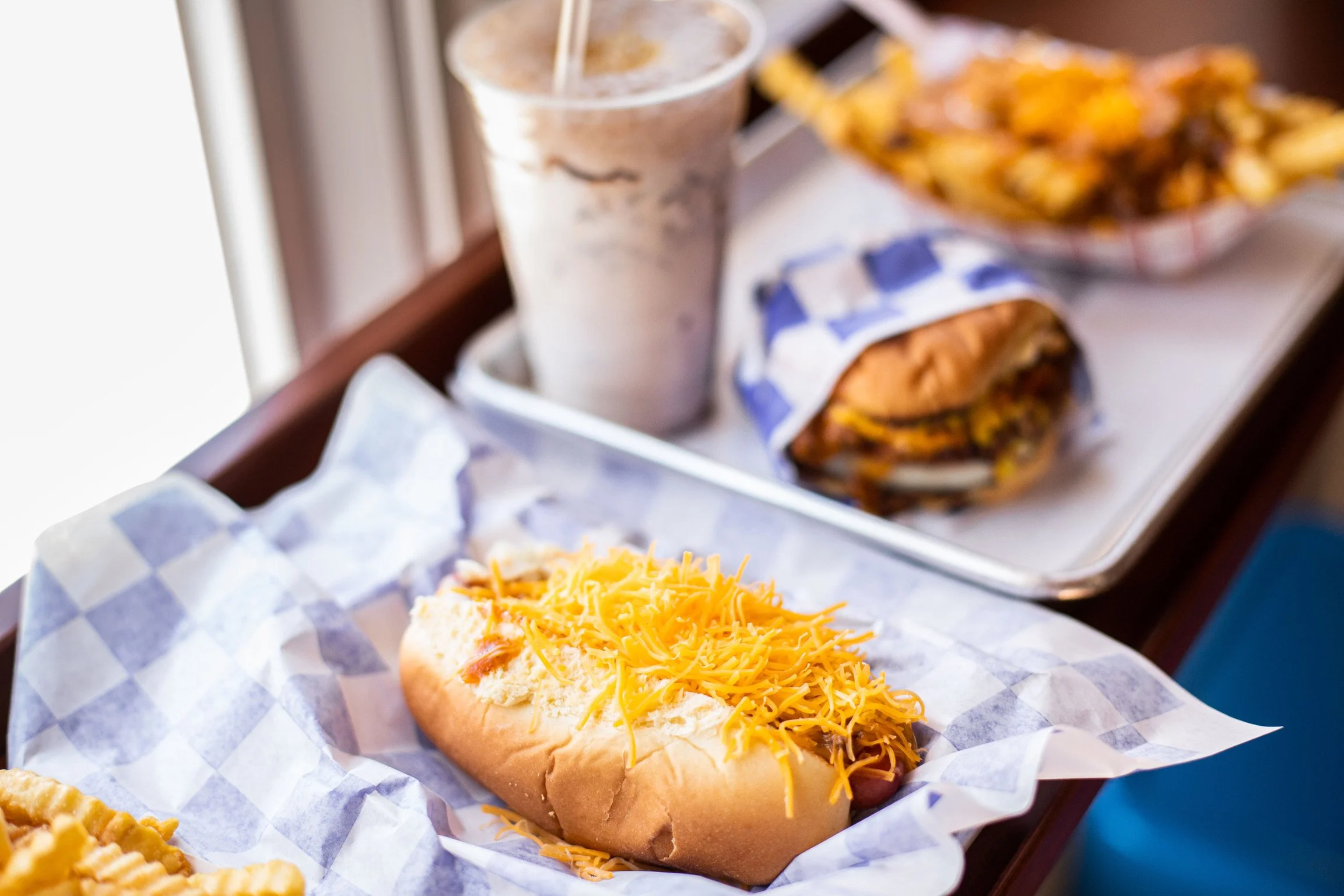 A hot dog with shredded cheese in a bun, served on a tray lined with checkered paper. In the background, there is a soda and a plate of curly fries.
