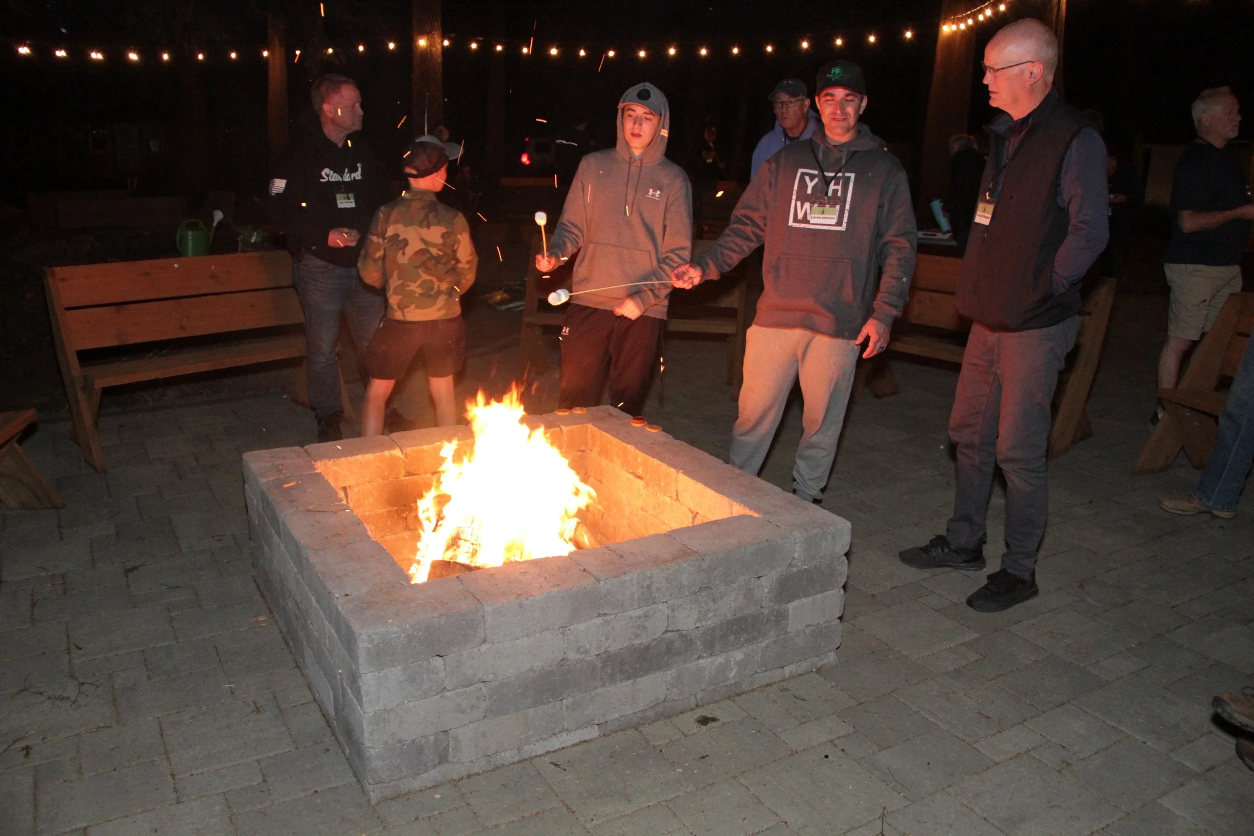 People gathered around a fire pit at night, some holding marshmallow sticks, with string lights overhead.